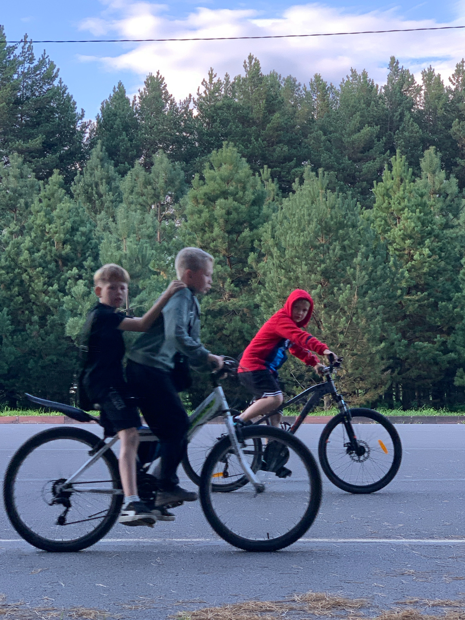 Three young boys are riding bicycles on a paved road in front of a dense forest. The boy on the left, wearing a black t-shirt and shorts, is being held onto by the boy in the middle, who is wearing a green hoodie and dark pants. The boy on the right, wearing a red hooded sweatshirt and black shorts, is riding his own bicycle. The sky above is blue with scattered clouds, and a power line is visible against the sky. The scene appears to be in Prokopyevsk, Russia.