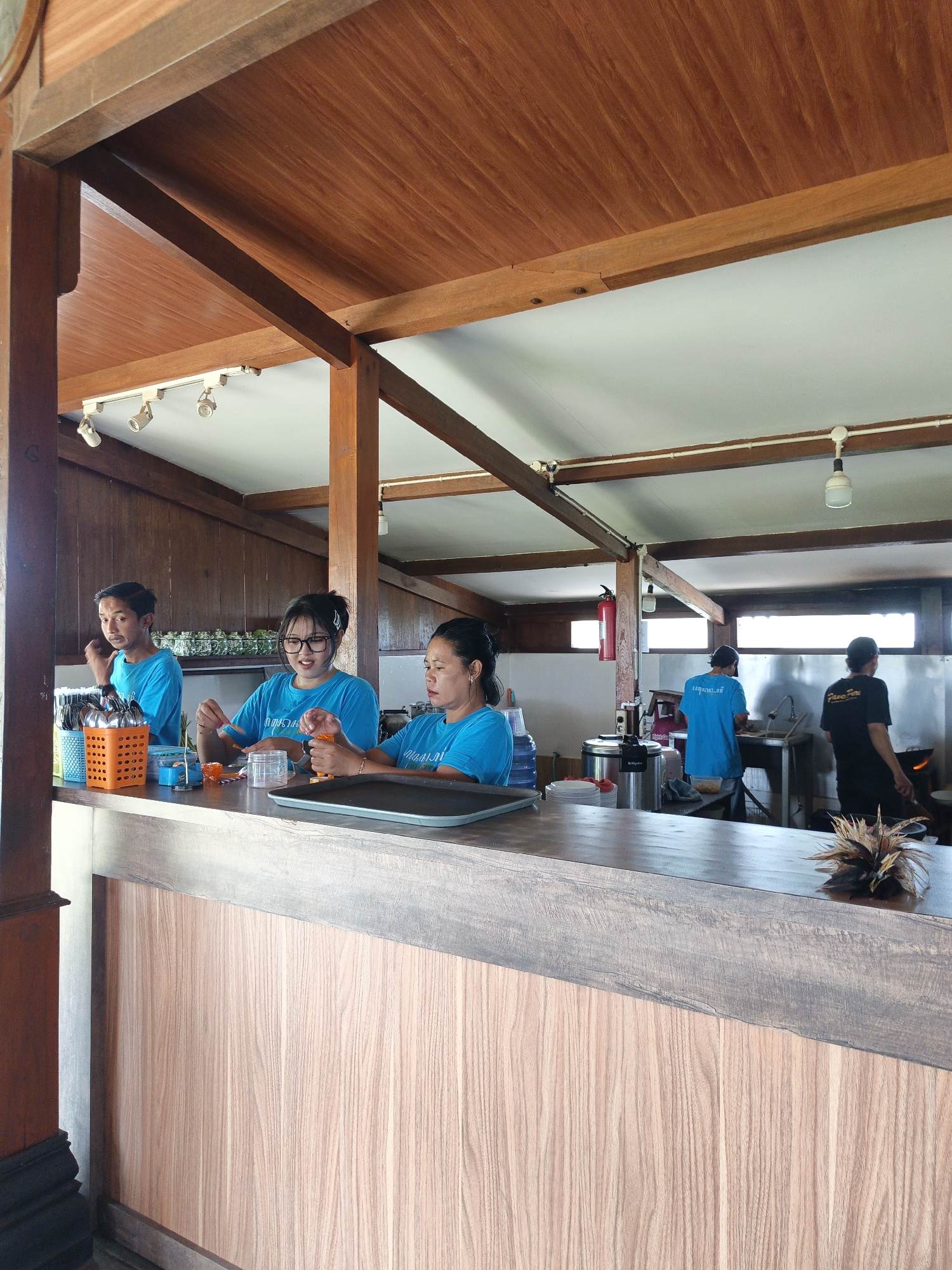 The image captures an interior scene, likely a cafe or restaurant, with a focus on a wooden counter and staff members. In the foreground, a light-colored wooden counter with a darker trim runs across the bottom half of the frame. Behind the counter, three individuals wearing bright blue t-shirts are visible.
On the left, a young man with short dark hair and a concerned expression is looking towards something out of frame. He is standing behind the counter. To his right, a young woman with glasses and dark hair tied back is holding an orange object in her right hand and a utensil in her left, appearing to be preparing something. Next to her, another woman with dark hair pulled back is also working, her hands obscured by the counter.
Further in the background, the scene opens up to a more functional area. Two men in blue shirts are visible near sinks and kitchen equipment. One man is facing away from the camera, working at a sink. Another man, wearing a black t-shirt, is also in the background, seemingly engaged in cooking activities near a stove.
The environment features a prominent wooden ceiling with visible beams, suggesting a rustic or tropical architectural style. White ceiling panels are visible between the beams. Lighting fixtures are attached to the ceiling. The walls behind the counter appear to be dark wood paneling, while the back area has windows that let in natural light. A red fire extinguisher is mounted on a wooden pillar in the background. The overall impression is of a busy service area within a establishment. This scene is located in Sekarsuli, Indonesia.