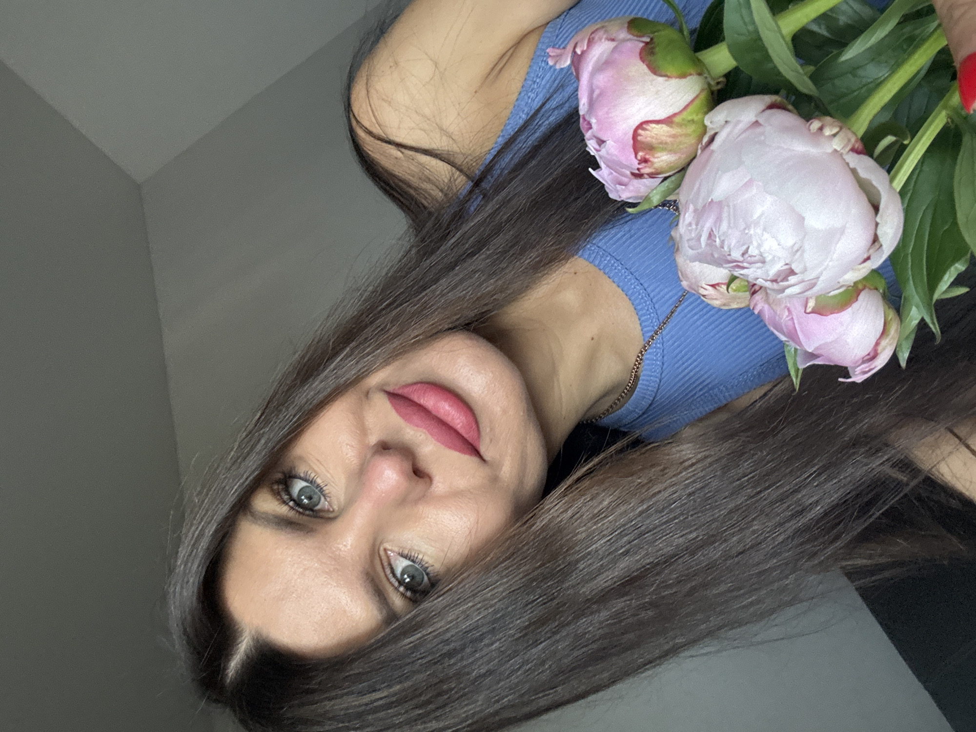 A woman with long, dark hair and blue eyes, wearing a blue top and a gold necklace, is holding a bouquet of pink peonies. Her lips are painted with a matte pink lipstick. The background is a plain, light gray wall. The image is a close-up shot, with the woman's face and the flowers filling most of the frame. The lighting appears to be natural daylight, coming from the left. There is no indication of the specific location within Chaikovsky, Russia, as the image only shows an indoor setting with a plain wall.