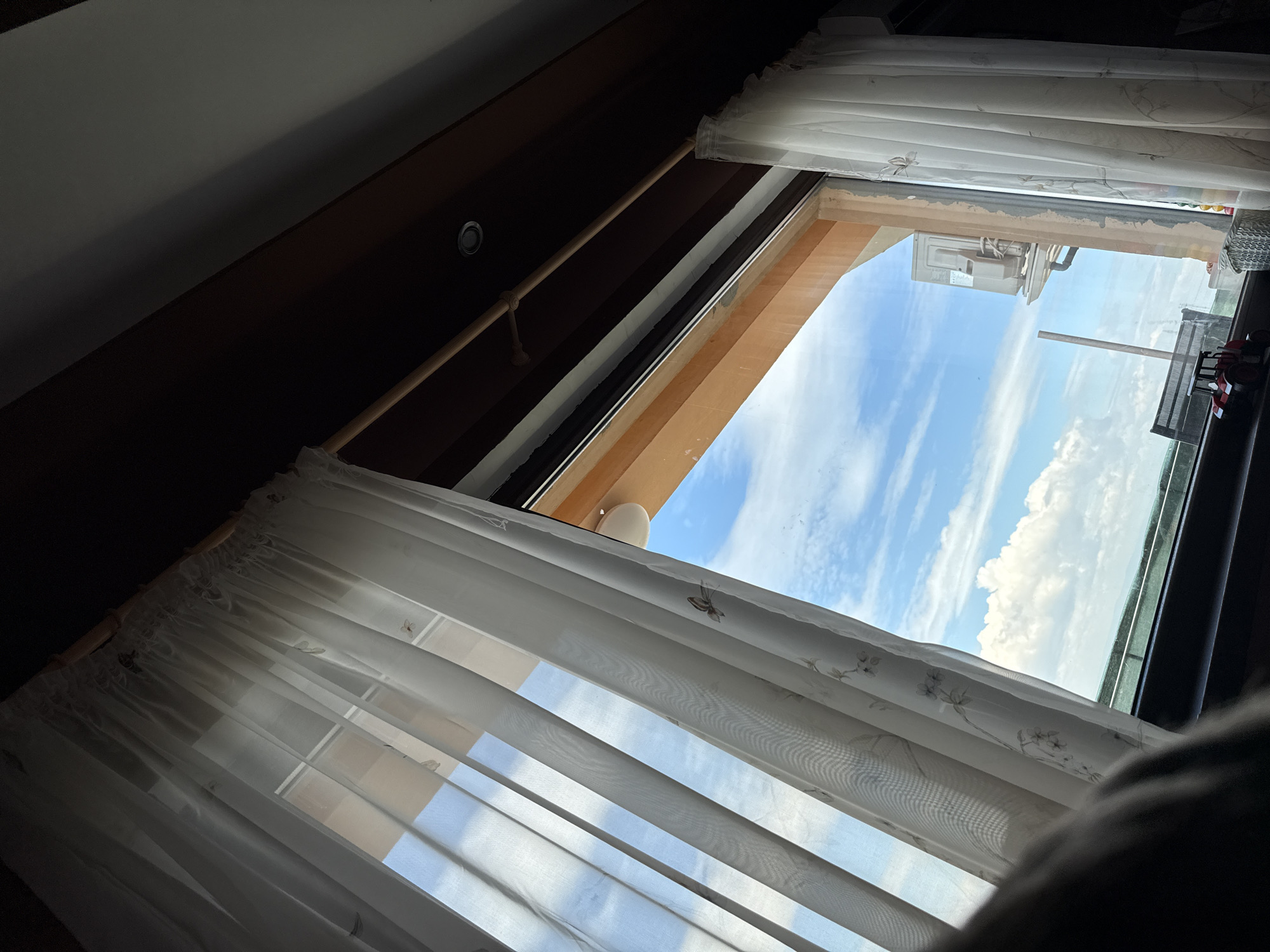 A view from inside a building, likely a residential space, shows a window partially covered by a white, sheer curtain. The window frame is visible, revealing an exterior view of a blue sky dotted with white clouds. An adjacent building is visible through the window. It is impossible to determine if this image was taken from the city of Sofia, Bulgaria without additional information.