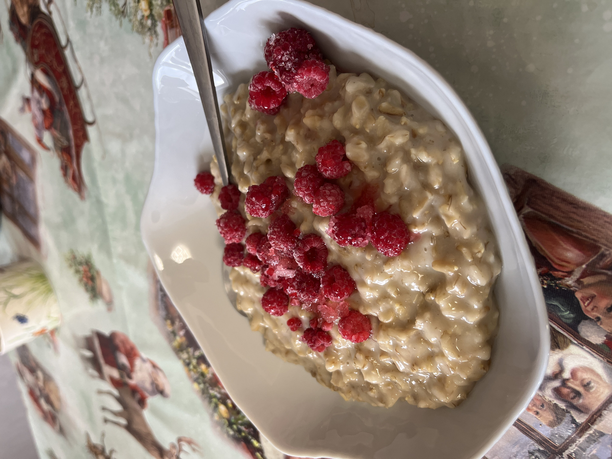 A top-down, slightly angled, medium shot captures a white, irregularly shaped bowl of oatmeal topped with a scattering of red raspberries. A silver spoon rests within the bowl. The bowl sits on a patterned tablecloth featuring images of people and deer. The scene is indoors, likely in a private residence, although the exact location within the city of Chaikovsky, Russia, cannot be determined from the image alone. The focus is on the food item, with the background providing context to the setting.