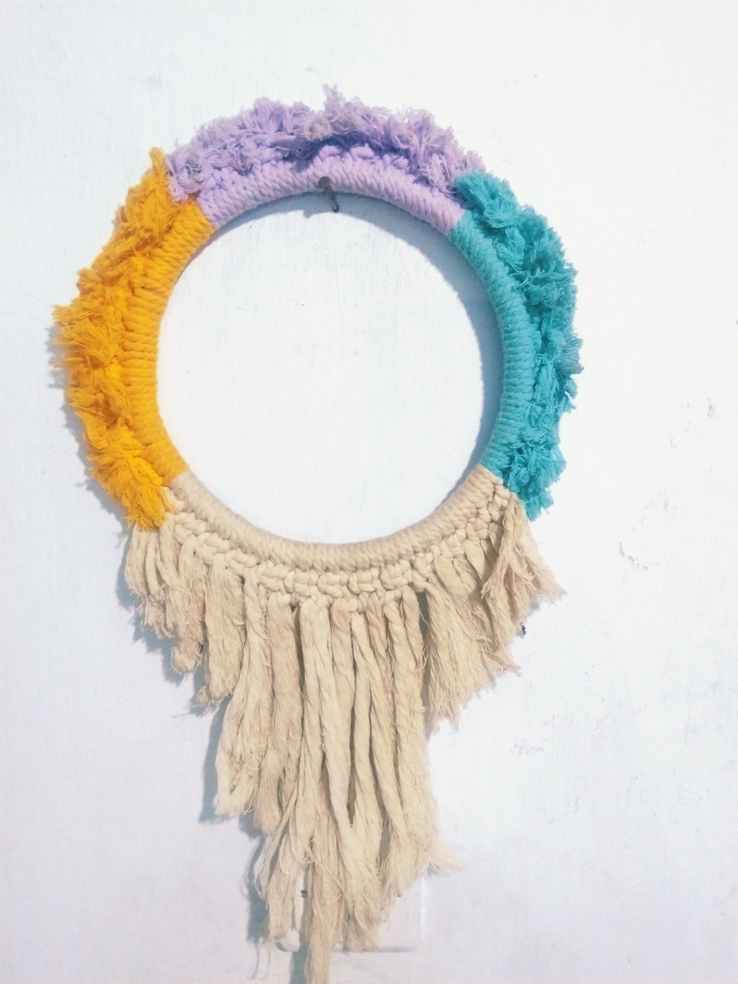 The image shows a wall decoration made of woven yarn, hanging on a white, textured wall. The decoration is circular in shape and is adorned with yarn in various colors: yellow, purple, turquoise, and cream. The cream yarn hangs down from the bottom of the circle, forming a fringe. There is a small nail or hook visible on the wall, just above the purple section of the yarn. The lighting suggests it is daytime. The scene does not provide any specific visual cues to determine its location in Tasikmalaya, Indonesia.