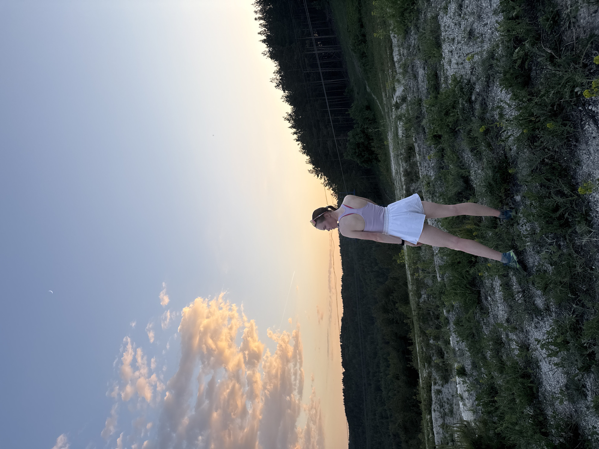 A woman in a pink tank top and white shorts stands on rocky, grassy terrain in an outdoor setting. The time of day appears to be dusk, judging by the sunset-colored sky filled with clouds. The background consists of a line of trees on the horizon. In the upper left of the image, there is a sliver of a crescent moon in the sky. A contrail left by a plane is visible above the clouds in the sky. The location is likely near Khvalynsk, Russia, given the landscape and available context.