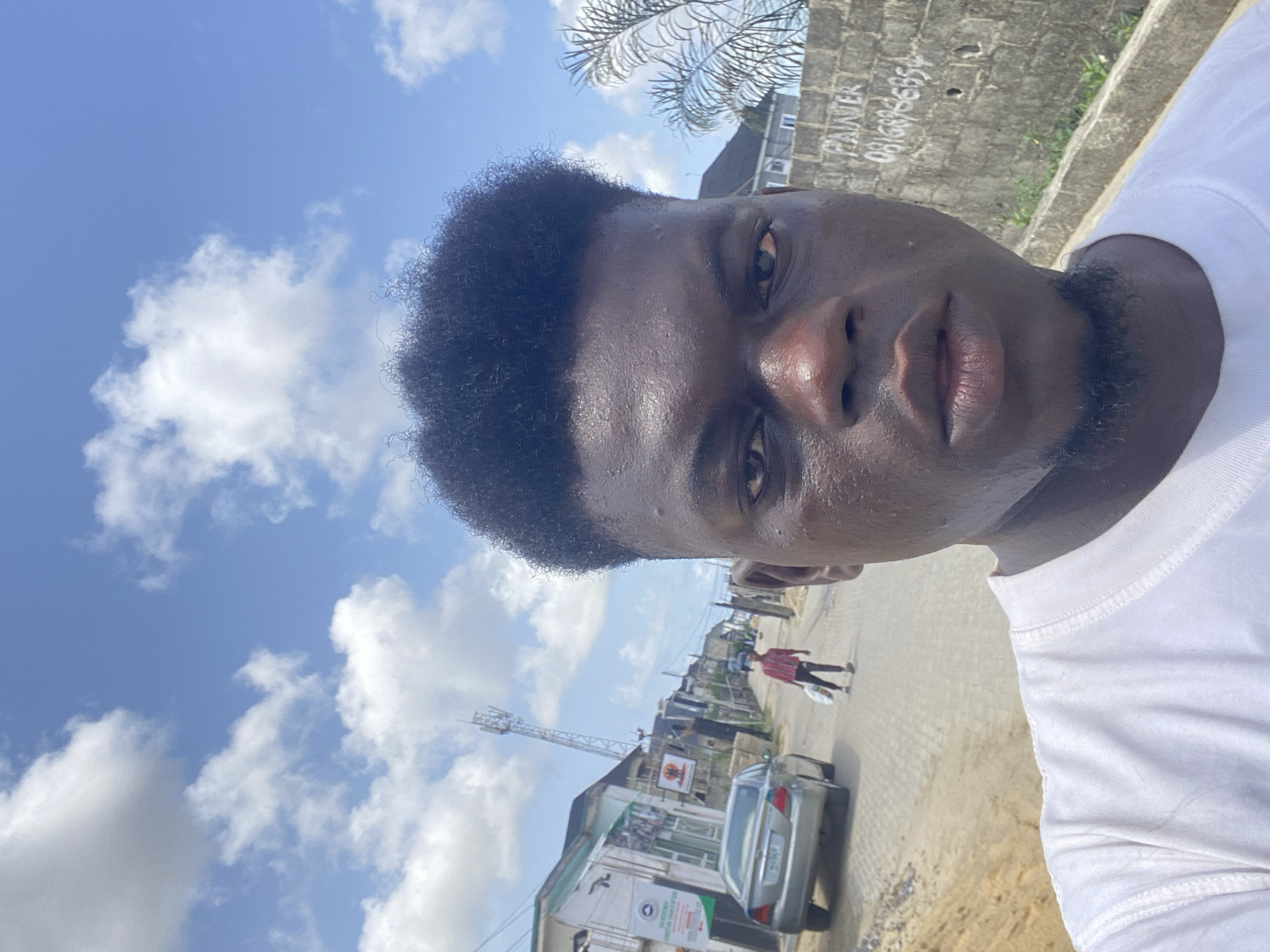 A selfie taken outdoors features a young Black man dominating the foreground, looking directly at the camera with a neutral to slightly serious expression. He has short, dark curly hair and visible stubble/beard, and his skin appears shiny, possibly from sweat. He is wearing a white T-shirt.
The background reveals a bright, sunny urban or semi-urban street scene under a clear blue sky dotted with scattered white clouds. A paved road stretches into the distance, where a person is seen walking away from the camera and a silver car is parked or driving. Low-rise buildings line the street, and a tall communication tower stands further back. Palm tree fronds are visible in the upper background.
Notable details include a weathered grey wall with the word "PAINTER" and the phone number "0816966654" spray-painted on it. Another sign, partially visible on a building, displays green and white elements. The overall impression is that of a warm, active day.