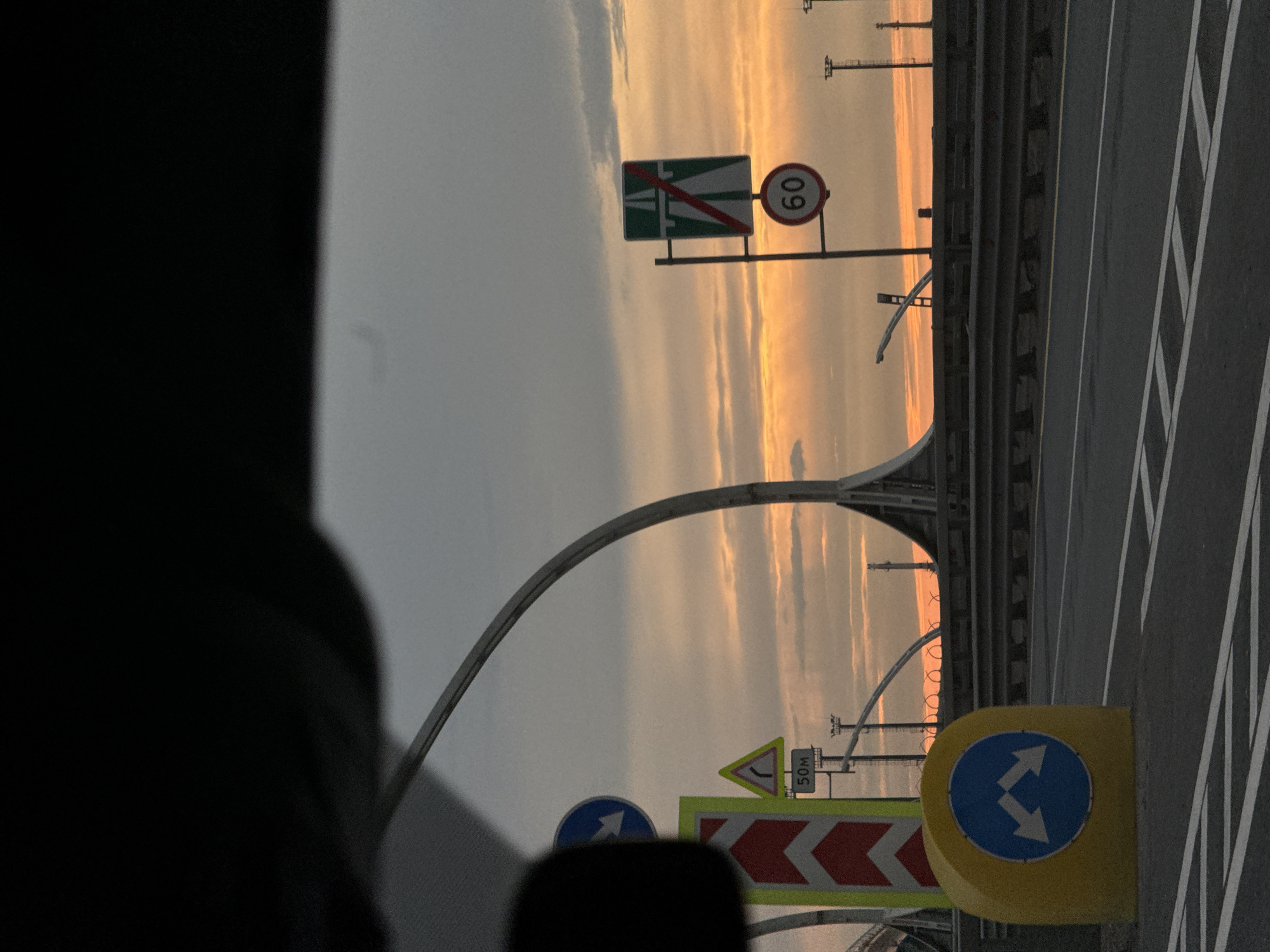 The image shows a road scene at sunset with several traffic signs. In the foreground, on the left, a yellow bollard displays a blue circular sign with two white arrows pointing left and right, indicating a bifurcation of the road ahead. Next to it, a tall, vertical sign with red and white chevron markings is visible. Further back, a triangular yellow sign with a black curved arrow indicating a sharp turn is mounted, with a smaller sign below it reading "50M". To the right of the road, a signpost holds two traffic signs: an upper green rectangular sign with white diagonal stripes indicating the end of a highway, and below it, a red circular sign with the number "60" signifying a speed limit of 60 kilometers per hour. The road itself is asphalt with white lane markings. In the background, a large, curved structure, likely part of a bridge or overpass, is prominent against a cloudy sky illuminated by the warm colors of the setting sun. This scene is likely located on Krestovsky Island in Saint Petersburg, Russia, possibly on the approach to or on the Krestovsky Bridge, given the presence of such modern traffic infrastructure and the distinctive sunset lighting. There are no people visible in the image.