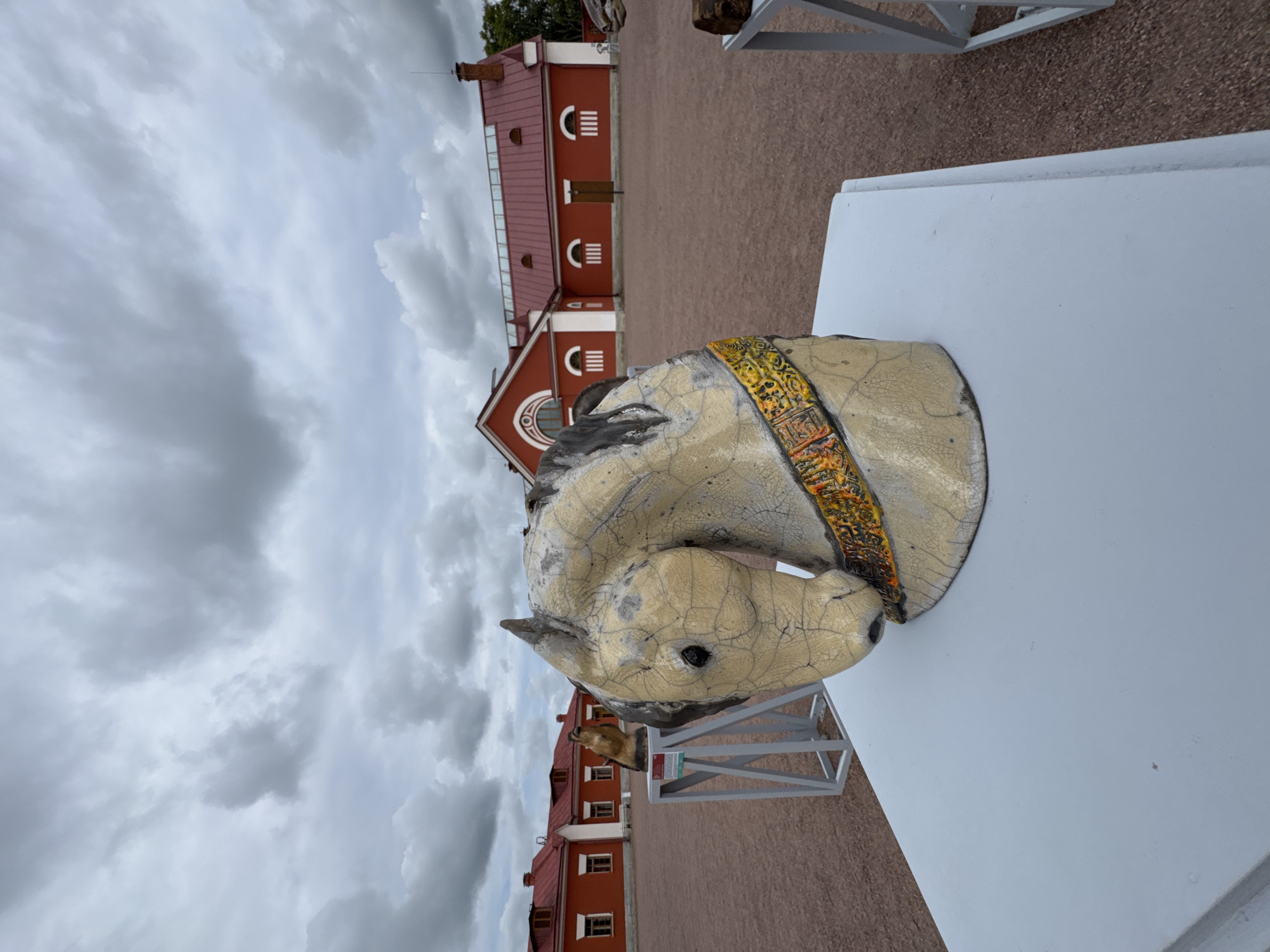 The image depicts an outdoor exhibition featuring horse head sculptures in the city of Pushkin, Russia, specifically at the Pavlovsk Palace. In the foreground, a cracked, beige ceramic horse head sculpture is displayed on a light gray pedestal. It has a dark mane and a decorative collar with yellow, orange, and black patterns. Behind it, another horse head sculpture, appearing to be made of a darker material, is placed on a gray metal stand with a white label. In the background, a long, red building with white trim and arched windows is visible, characteristic of the architecture found in Pushkin. The ground is covered with reddish-brown gravel. The sky is overcast with gray clouds, suggesting a cool, possibly damp day.