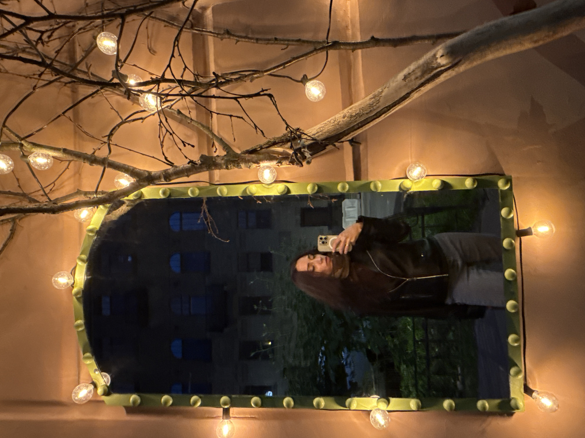 A young woman with long, dark hair stands in front of a large, ornate mirror, taking a selfie with her phone. She is wearing a black leather jacket, a light brown scarf, and grey pants. The mirror is framed with a green border adorned with a string of small, glowing lights. The background reflected in the mirror shows a dark building with windows and some greenery. To the right of the mirror, bare tree branches are intertwined with more strings of lights. The overall scene is dimly lit, with the warm glow of the lights creating a cozy atmosphere. The image appears to be taken outdoors in Saint Petersburg, Russia, given the architectural style of the building in the reflection.