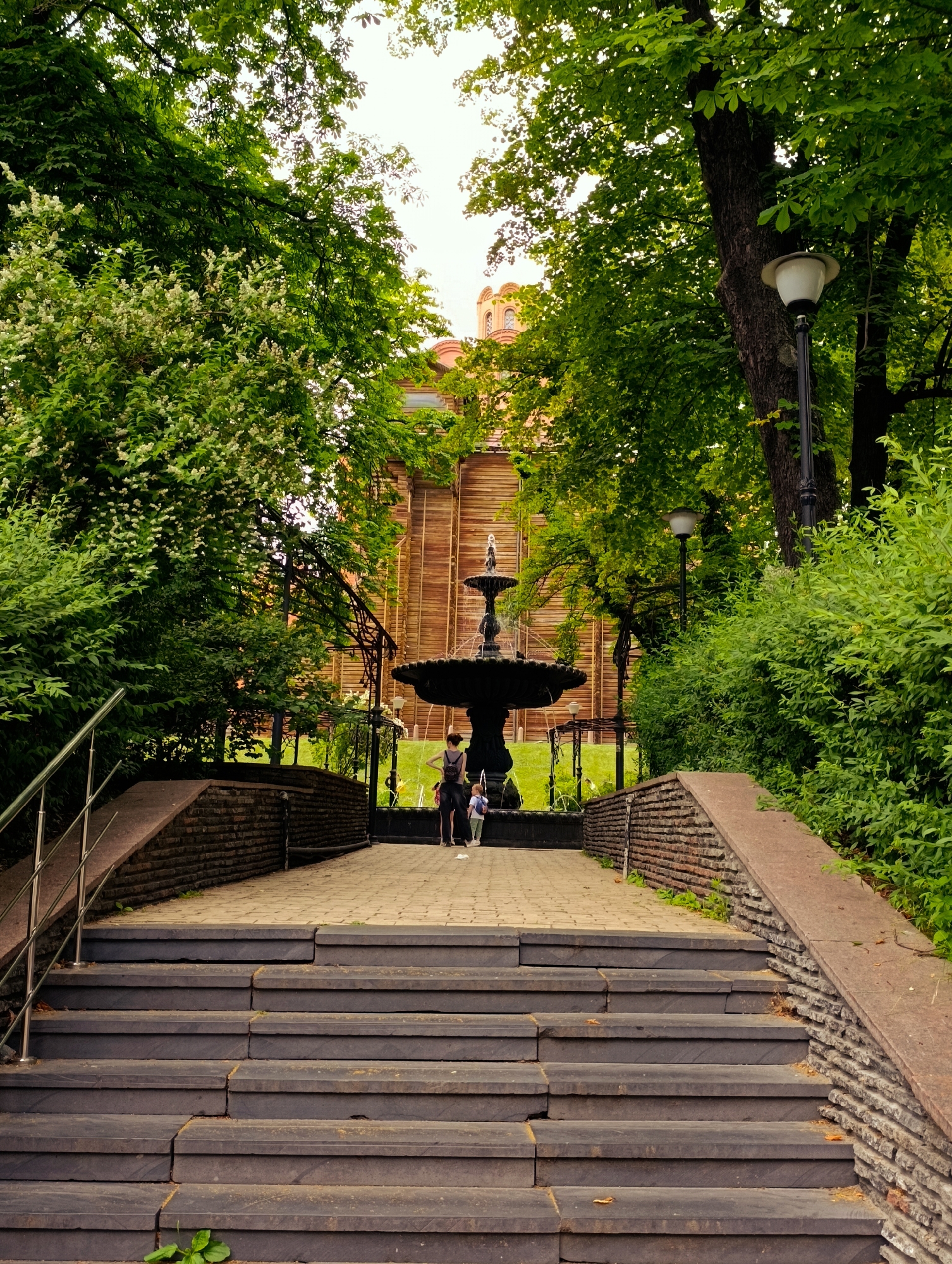 The image shows a stone staircase leading up to a pathway in a park. On the pathway, a black multi-tiered fountain is visible with water spraying from it. Two people, one adult and one child, are standing near the fountain, looking at it. Behind the fountain, a building with wooden paneling is visible. Trees and bushes surround the scene, with a lamppost to the right. The scene is set outdoors, in a park-like environment. This park is located in Kyiv, Ukraine, specifically in the Shevchenko district.