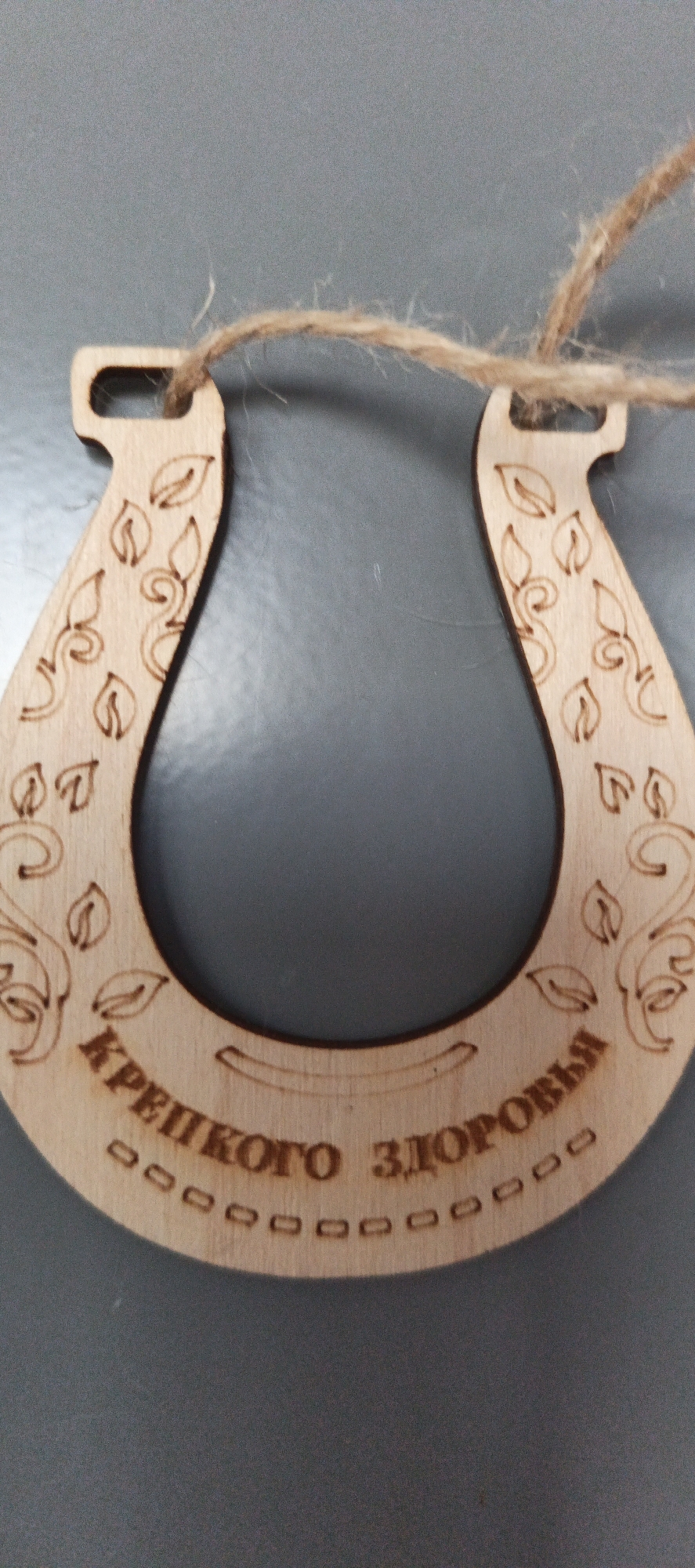 A wooden horseshoe charm hangs on a gray surface by a piece of twine. The charm is carved with decorative leaf and vine patterns on its upper arms. Along the bottom curve of the horseshoe, the Russian text "КРЕПКОГО ЗДОРОВЬЯ" (KREPKOVGO ZDOROVYA), which translates to "Good Health," is engraved, with a line of small, hollow squares beneath it. The horseshoe is positioned vertically, with its open end facing downwards. The twine loops through two small holes at the top of the horseshoe. The background is a plain, solid gray. There are no people or other objects visible in the image. This object is likely a souvenir or gift. The location of Tikhvin, Russia is not depicted in this image.