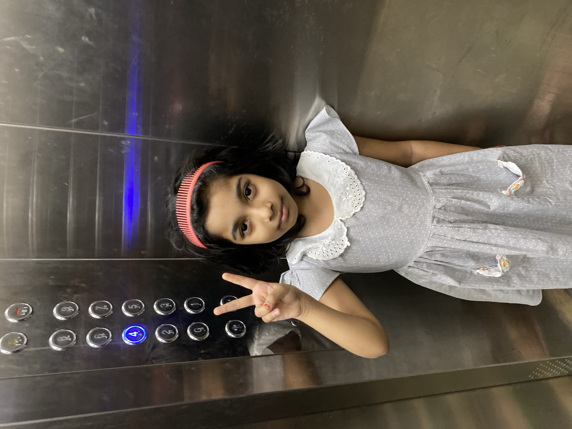 A young girl is featured inside an elevator in Dhaka, Bangladesh, captured in a portrait-style shot that appears rotated 90 degrees clockwise. She has dark hair styled with a bright pink, comb-like headband and is wearing a light-colored dress with fine stripes or dots, accented by a distinctive white, scalloped lace collar. She looks directly at the camera with a neutral to slightly smiling expression.
With her right hand, the girl makes a peace sign, holding it near the elevator's control panel. The panel, set into the metallic elevator wall, displays a grid of circular buttons. The button for "4" is prominently illuminated with a bright blue light, indicating an active selection or current floor. Other visible buttons include numbers like 1, 2, 5, 6, 7, 8, 9, 10, and a "G" button, presumably for the Ground floor, along with other functional symbols. The elevator interior consists of brushed metal walls that show minor scuffs and reflections. The scene depicts a casual moment inside an everyday urban setting.