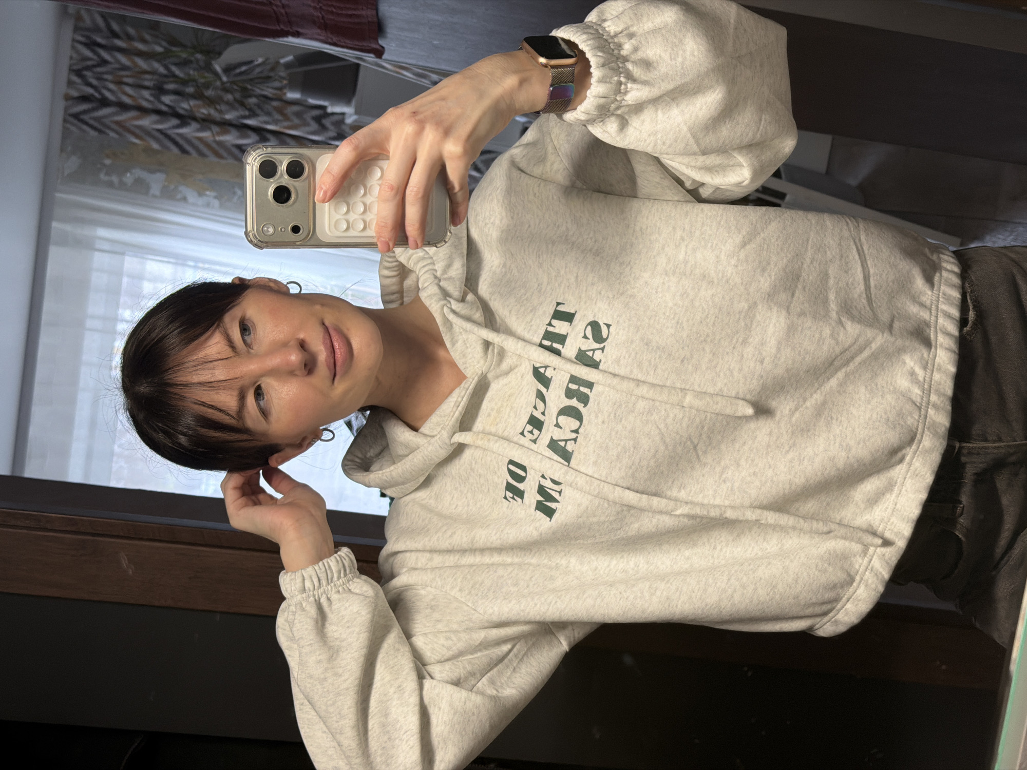 A person with short, dark hair and bangs is taking a selfie using a smartphone. The phone is held in their right hand, which also has a smartwatch. They are wearing a light gray hoodie with dark green text. The background includes a window with a white curtain and a dark brown shelf or countertop. The reflection in the mirror shows part of a room with a dark object. The specific location in the city of Perm, Russia cannot be determined from the image.