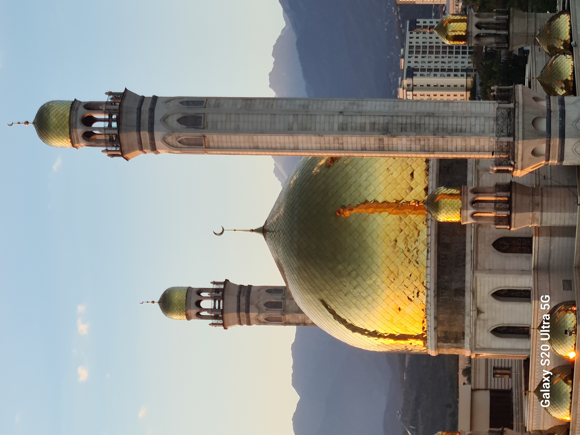 The image shows the Zenkov Cathedral in Almaty, Kazakhstan, at sunset. The cathedral is a Russian Orthodox church made entirely of wood and is one of the tallest wooden buildings in the world. It is located in Panfilov Park, a popular tourist destination in the city. The cathedral is painted in bright colors and has a golden dome. The sun is setting behind the mountains, casting a warm glow on the cathedral and the surrounding park. There are no people visible in the image.