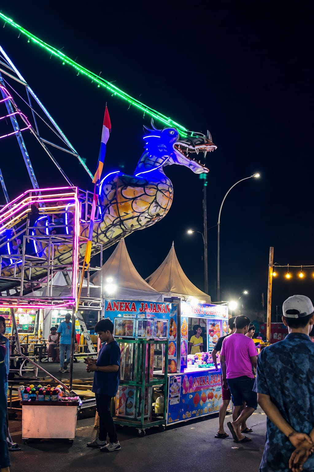 At night, a brightly lit amusement park in the City of Medan, Indonesia, features a large, inflatable dragon sculpture with blue and yellow accents, illuminated by neon lights. The dragon is positioned next to a Ferris wheel, with green and pink lights tracing its structure. Below, several food stalls are set up, displaying various snacks and drinks, with "ANEKA JAJANAN" (meaning "various snacks" in Indonesian) prominently advertised. People are seen walking around, purchasing food, and interacting with stall vendors. The atmosphere is lively and festive, with the dark night sky providing a backdrop to the colorful lights and attractions.