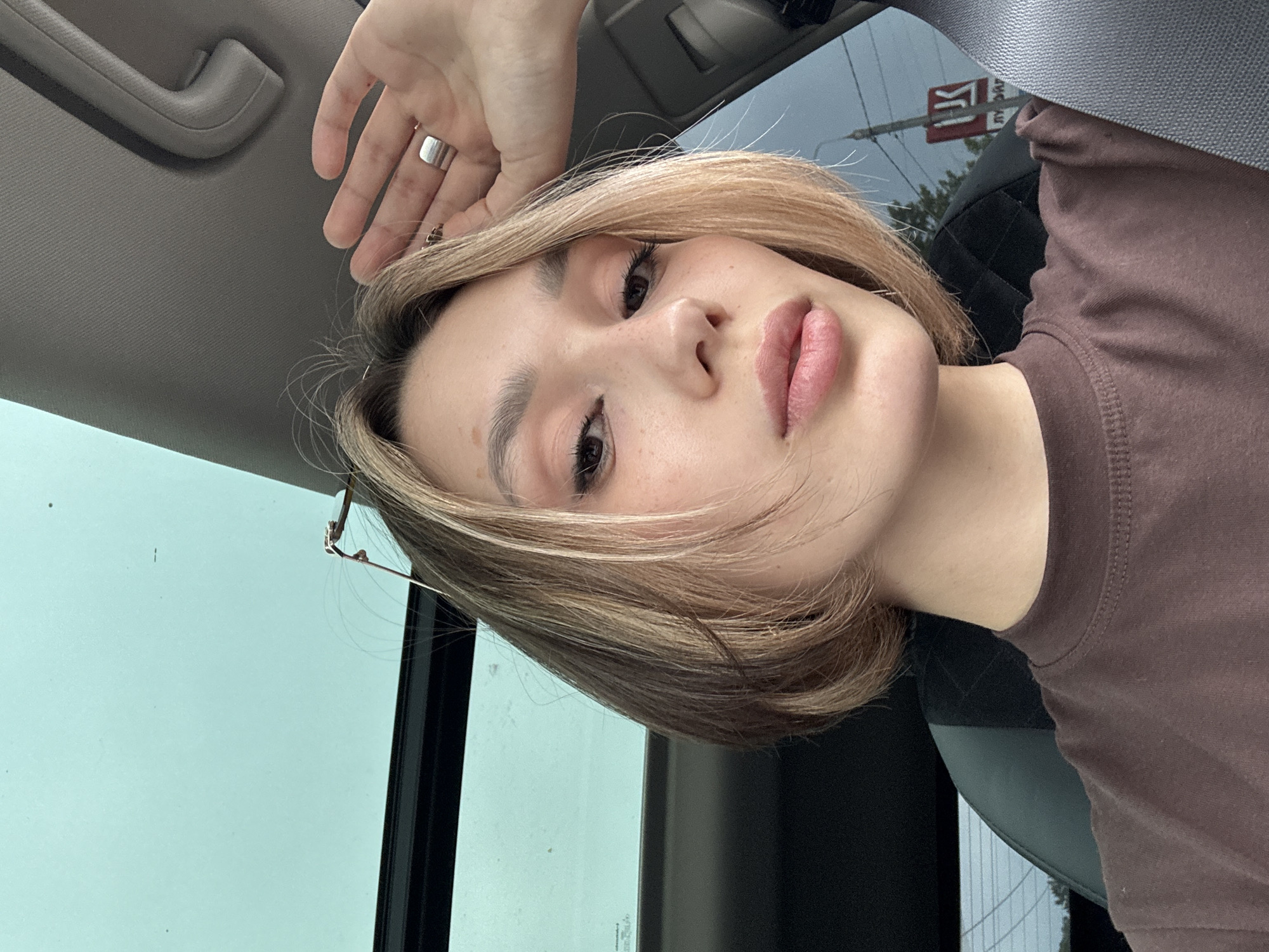 A young woman with short, blonde hair is taking a selfie in a car. She is wearing a brown t-shirt and a silver ring on her right hand, which is raised to her forehead. Her eyes are closed and her lips are slightly parted. The car has a sunroof, and the interior is visible, including a seatbelt and part of the dashboard. Outside the car, trees and a red sign with Cyrillic lettering are visible through the window. The scene is set in an urban environment, as indicated by the sign and the surrounding buildings.