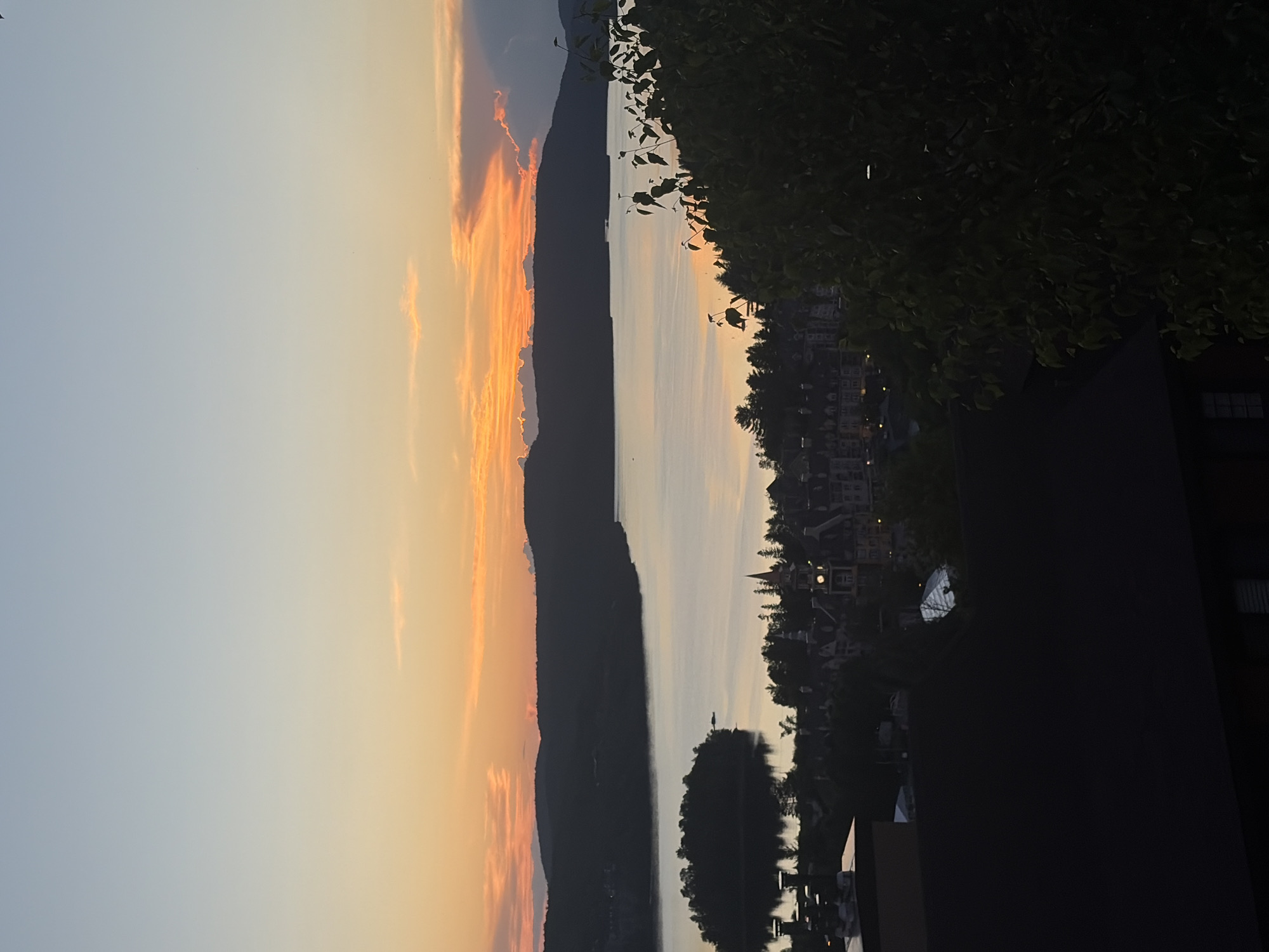 The image shows a panoramic view of Mont-Tremblant, Canada, during a sunset. The sky is a gradient of soft blues and oranges, with scattered clouds tinged by the setting sun. In the foreground, dark silhouettes of trees and buildings obscure the lower portion of the scene.
The mid-ground features a body of water, likely Lac Tremblant, reflecting the warm colors of the sky. Across the lake, a cluster of buildings forms a village, characterized by its distinctive architecture with steeply pitched roofs and spires, reminiscent of European alpine villages. A prominent clock tower stands out among the structures. An island with dense vegetation is visible in the middle of the lake, closer to the village.
The background is dominated by the dark, undulating silhouette of the Mont Tremblant mountains, framing the tranquil waters and the picturesque village. The overall impression is one of serene natural beauty combined with the charm of a well-preserved resort town. There are no people visible in the image.