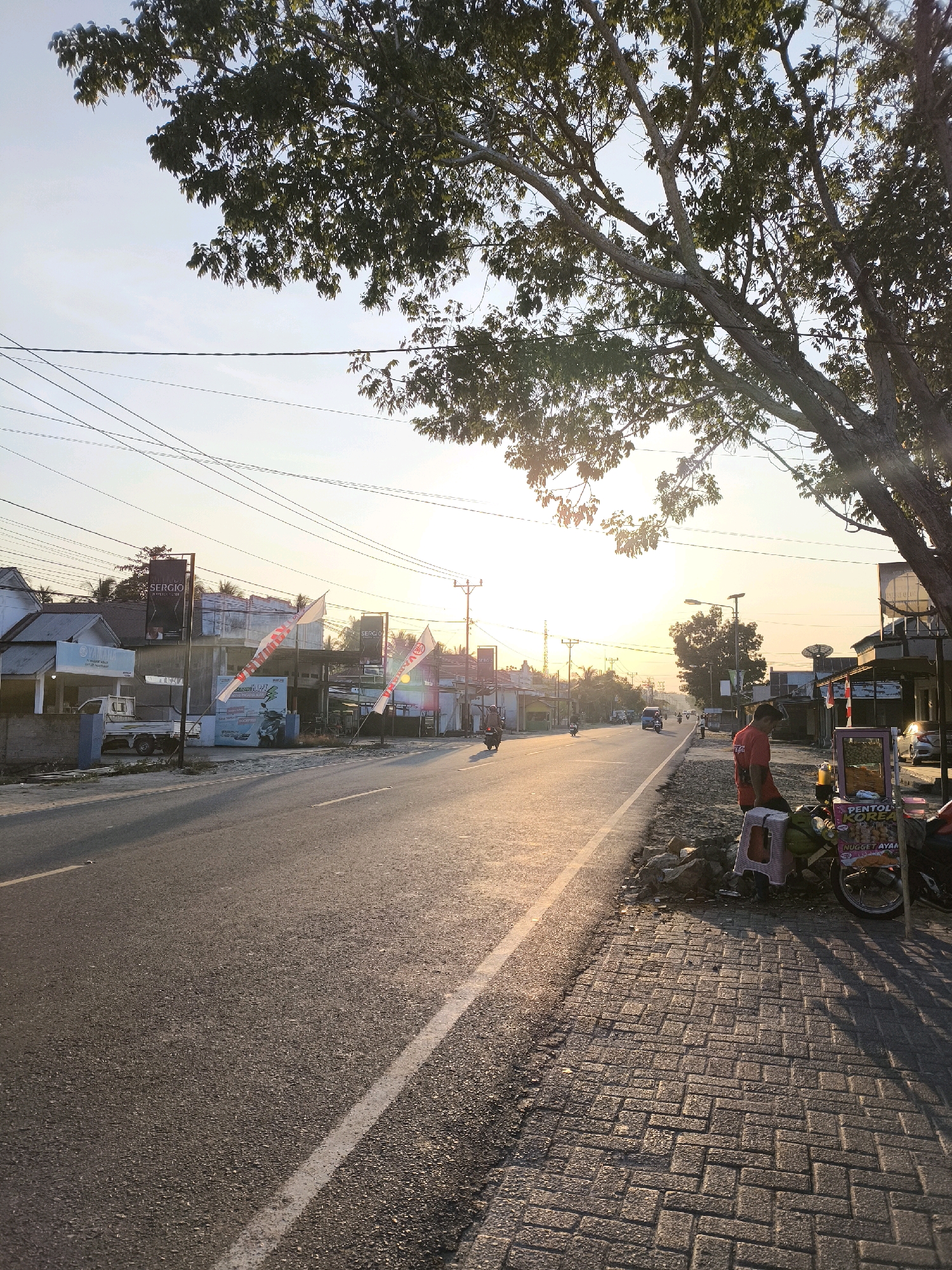 This is a photograph taken at street level in Tenilo, Indonesia, during what appears to be sunrise or sunset. The image captures a paved road running horizontally across the frame, with a wide sidewalk made of interlocking bricks on the right side. A large tree with dense foliage overhangs the scene from the upper right corner, casting shadows.
On the left side of the road, there are several buildings that appear to be shops or commercial establishments. One building prominently displays a sign that reads "SERGIO MOTORCYCLES." There are also flags and banners, some with "YAMAHA" logos. A light-colored truck is parked in front of one of the buildings. Overhead, a network of power lines crisscrosses the sky.
On the right sidewalk, a vendor is operating a small cart selling food items, with signs indicating "PENTOL KOREA" and "NUGGET AYAM." A person wearing a red shirt is standing next to the cart. A motorcycle is parked near the cart, and other motorcycles and a few cars can be seen further down the road. The sun is low on the horizon, creating a warm, golden light that illuminates the scene and casts long shadows. The overall environment suggests a typical street scene in a town or city in Indonesia.
