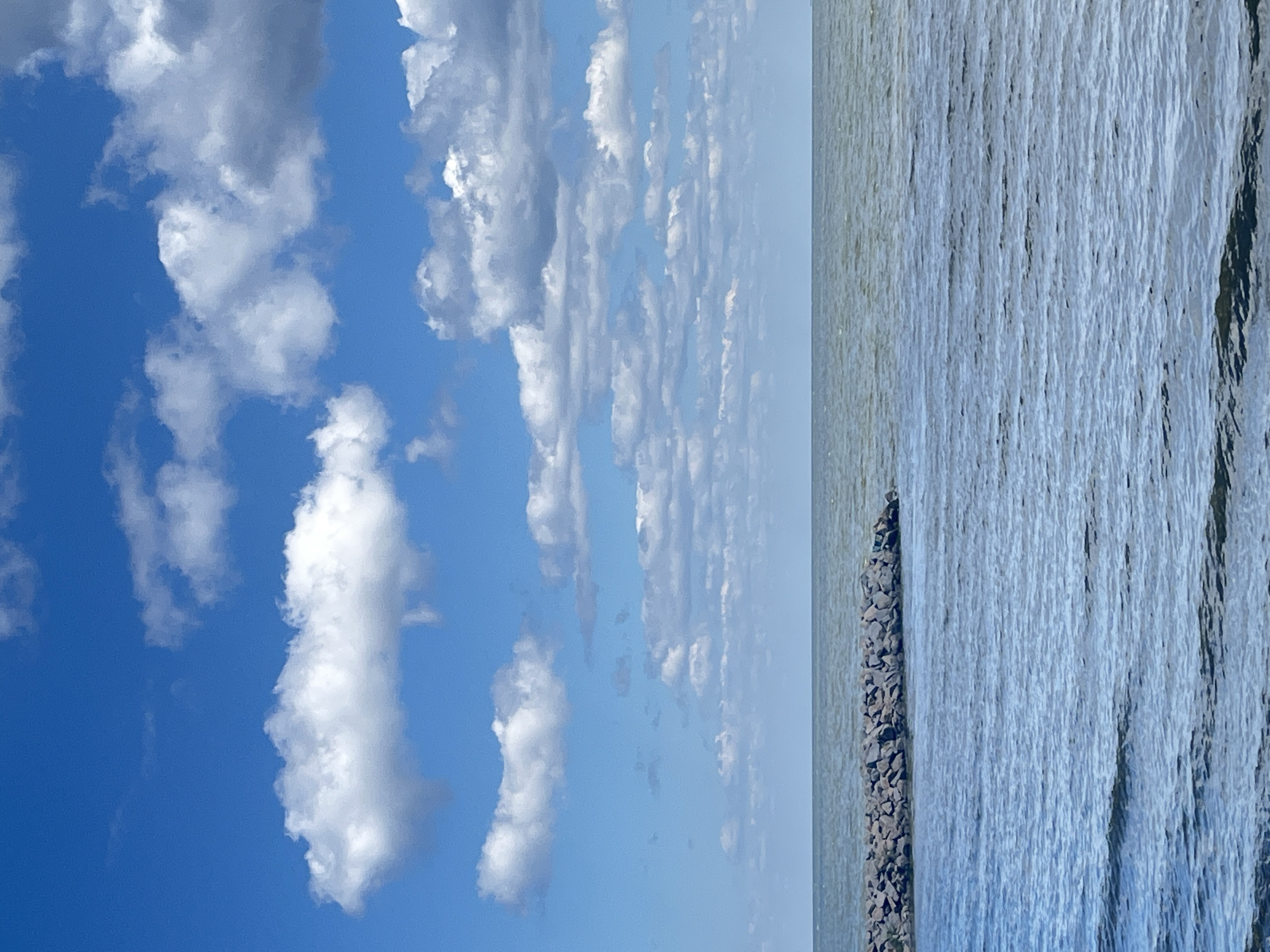 The image shows a body of water under a blue sky with scattered white clouds. In the foreground, the water has small waves, reflecting the sky. A dark stone breakwater juts out into the water from the left side of the frame, extending horizontally across the lower portion of the image. The horizon line, where the water meets the sky, is visible in the distance. The sky is a bright blue with large, fluffy white clouds in various sizes and densities, with some appearing higher and more distant than others. The scene appears to be an outdoor, natural environment, possibly a lake or sea, with no signs of urban development or people. This image is not located in Novi Petrivtsi, Ukraine; it depicts a generic seascape.
