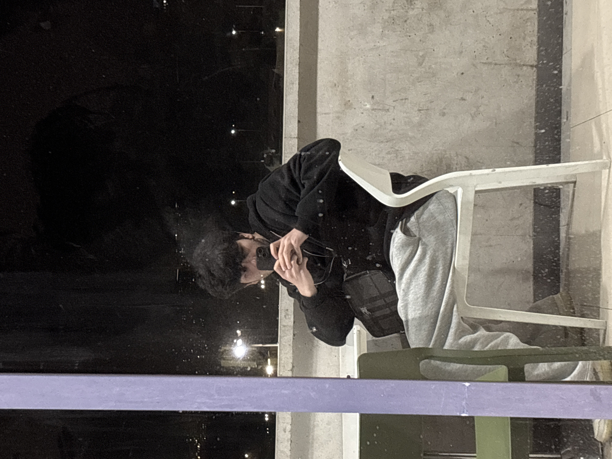 A person is reflected in a window. The person has dark hair and wears a black jacket and gray sweatpants. They hold a cell phone. The reflection also shows a white chair and a lavender-colored railing. The window reflects city lights at night. The image shows a scene reflected in a window in Yerevan, Armenia.