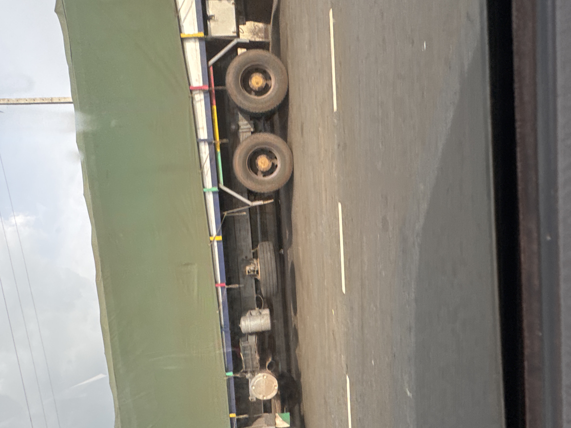 From an interior perspective, the scene depicts a portion of a moving truck, with its green canvas cover, tires, and undercarriage visible. The truck is traveling on a paved road with a painted white lane marker. Outside, the sky appears overcast. The location is Umuoye, Nigeria.