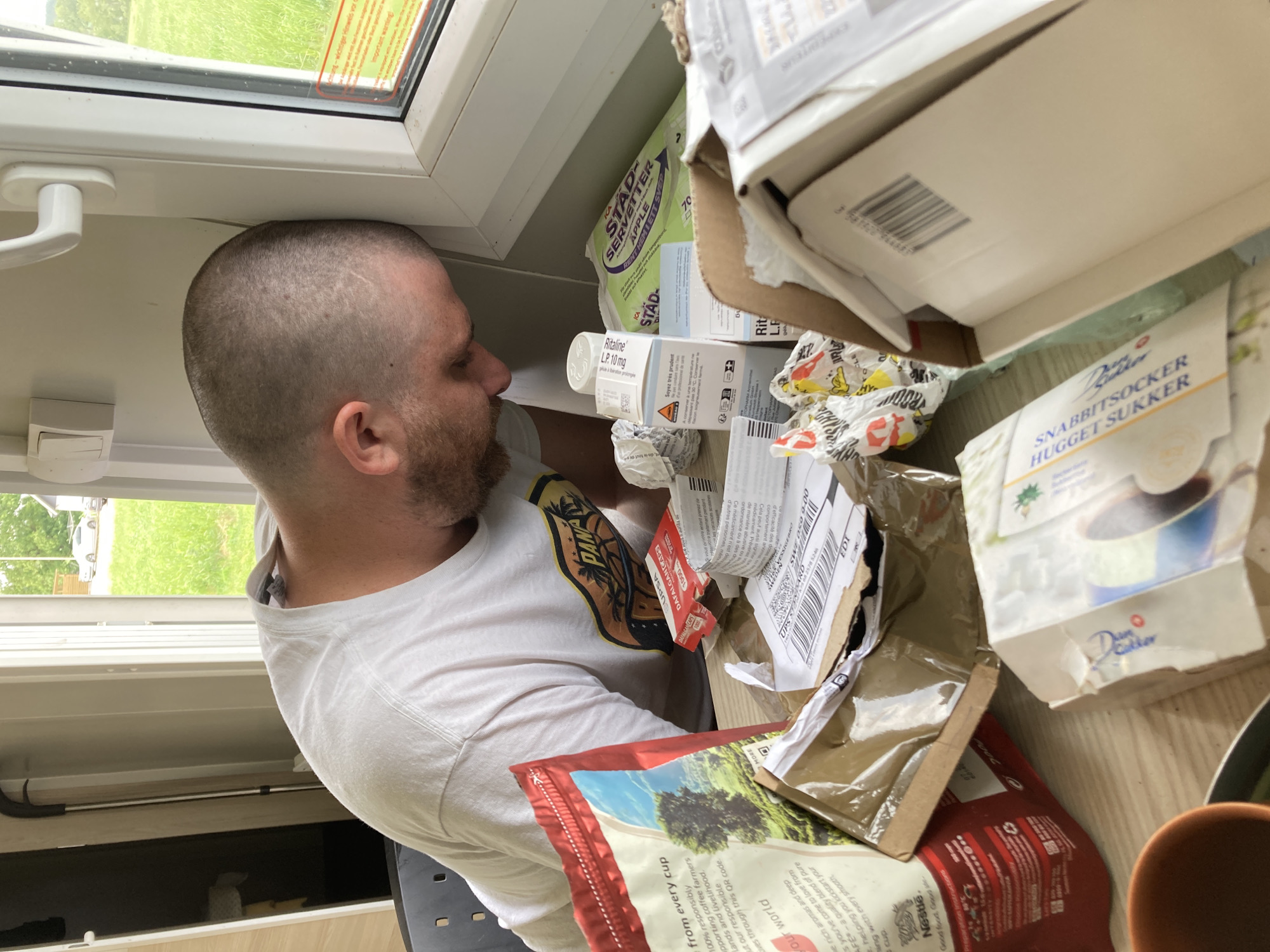 A man with short, buzzed hair and a beard is depicted in an indoor setting, likely in Vessigebro, Sweden, near a window. He is wearing a white short-sleeved t-shirt with a graphic design that includes the word "PANA". His gaze is directed downwards or towards a cluttered surface in front of him, suggesting he is examining or reaching for items.
The area next to him is densely packed with various packaged goods and boxes. Visible items include a box of "Ritaline L.P. 10 mg" (a medication), a package labeled "STAD-SERVETTER APPLE" (likely napkins or paper towels), and another package for "SNABBITSOCKER HUGGET SUKKER" (sugar cubes) from the brand "Dansukker". Other packaging includes a red bag with text related to "Nestle" and "100% responsibly supporting coffee farmers," and a red box partially visible with "UPSA DAFALGAN." A shipping label fragment shows "UPS STO 17163 VESSIGE MANG SWE OD 000," confirming the Vessigebro, Sweden location. The word "ICA" is also visible on some packaging.
The background features a window looking out onto a bright, green outdoor area with a white vehicle partially visible, indicating daytime with good natural light. The indoor surfaces are light-colored, possibly white or light wood. The scene portrays a moment of interaction with a collection of household or personal items on a shelf or counter.