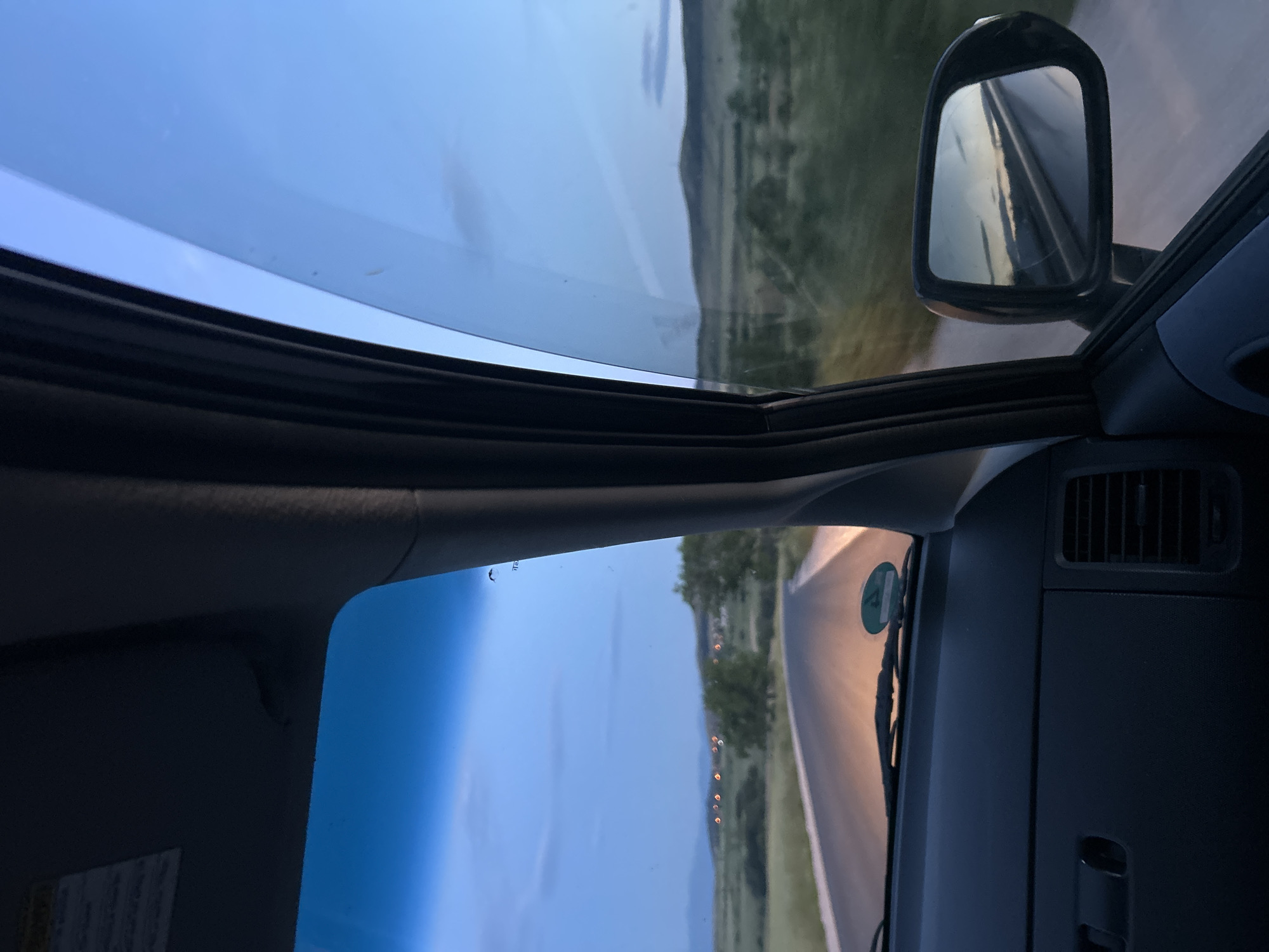 The image shows the view from inside a car driving on a road. The landscape is visible through the windshield and sunroof, featuring fields and a distant mountain. The side mirror reflects the road behind the car. The interior of the car is visible, including the dashboard and air vents. A roadworthiness sticker is on the inside of the windshield. The exact location in Ovchepoltsi, Bulgaria cannot be determined from this image.