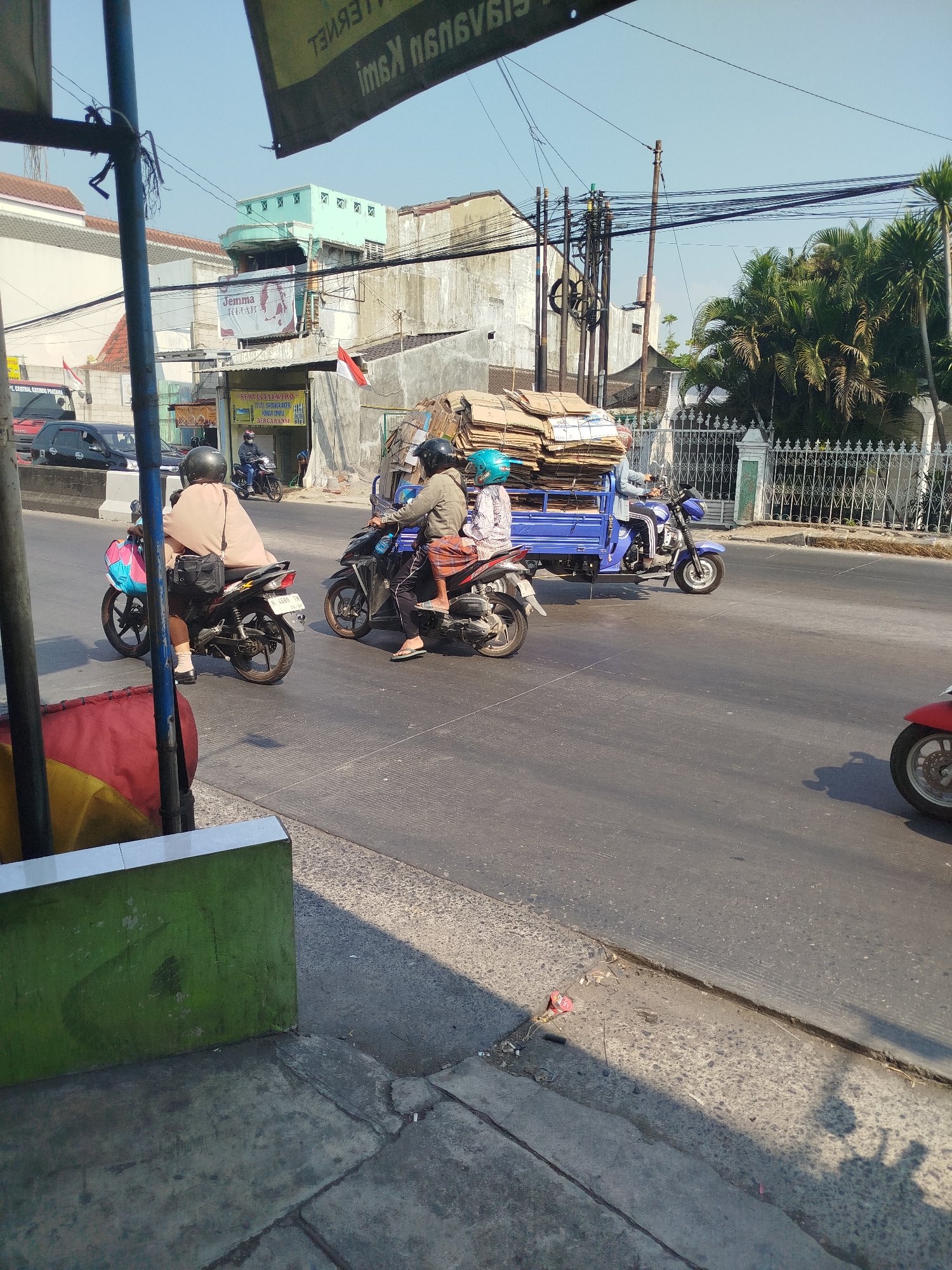 In Mranggen, Indonesia, a street scene unfolds under a clear blue sky. In the foreground, a green wall with a white tile trim borders a paved walkway. The street itself is paved and shows signs of wear. Several motorcycles are in motion. One motorcycle, ridden by a person wearing a light-colored long-sleeved shirt and shorts, moves from left to right. Another motorcycle, carrying two individuals, is also in transit, with the rider wearing a helmet and the passenger a blue helmet and a patterned top. Behind them, a blue three-wheeled vehicle is loaded with stacks of cardboard boxes, suggesting a delivery or transport activity. The vehicle is operated by a person wearing a helmet. In the background, buildings with varying architectural styles, including some with tiled roofs and a light green facade, are visible. Power lines crisscross the sky, supported by utility poles. Palm trees and other greenery add to the tropical environment. The overall impression is one of everyday life and commerce in a bustling Indonesian town.