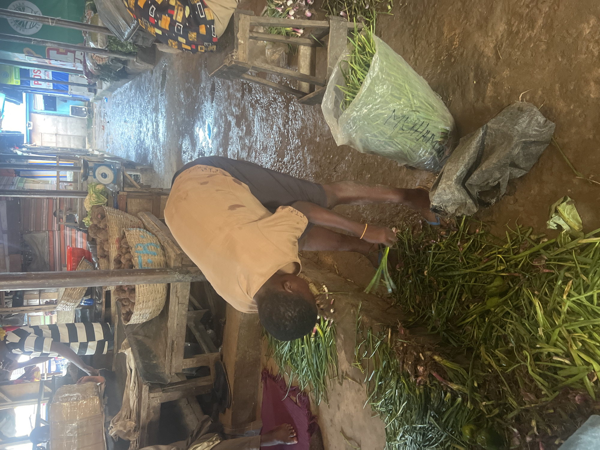 The image depicts a scene within a marketplace in Oko Amakom, Nigeria. In the foreground, a person is bent over, sorting through a pile of green onions and what appear to be red onions on the muddy ground. The person is wearing a light-colored t-shirt and dark shorts, with a yellow bracelet on their left wrist. To the right of the person, a clear plastic bag filled with more green onions is visible, alongside a dark grey plastic bag. Further to the right, a wooden stool stands beside a stall displaying produce, including bunches of onions. In the background, a pathway between market stalls is flooded with water, reflecting the overhead structures. On the left side of the image, another person is standing near market stalls piled with goods, including what looks like boxed items wrapped in plastic. Baskets filled with produce, possibly yams, are also visible on elevated wooden surfaces. The overall environment is that of a busy, albeit somewhat disorganized, outdoor market.
