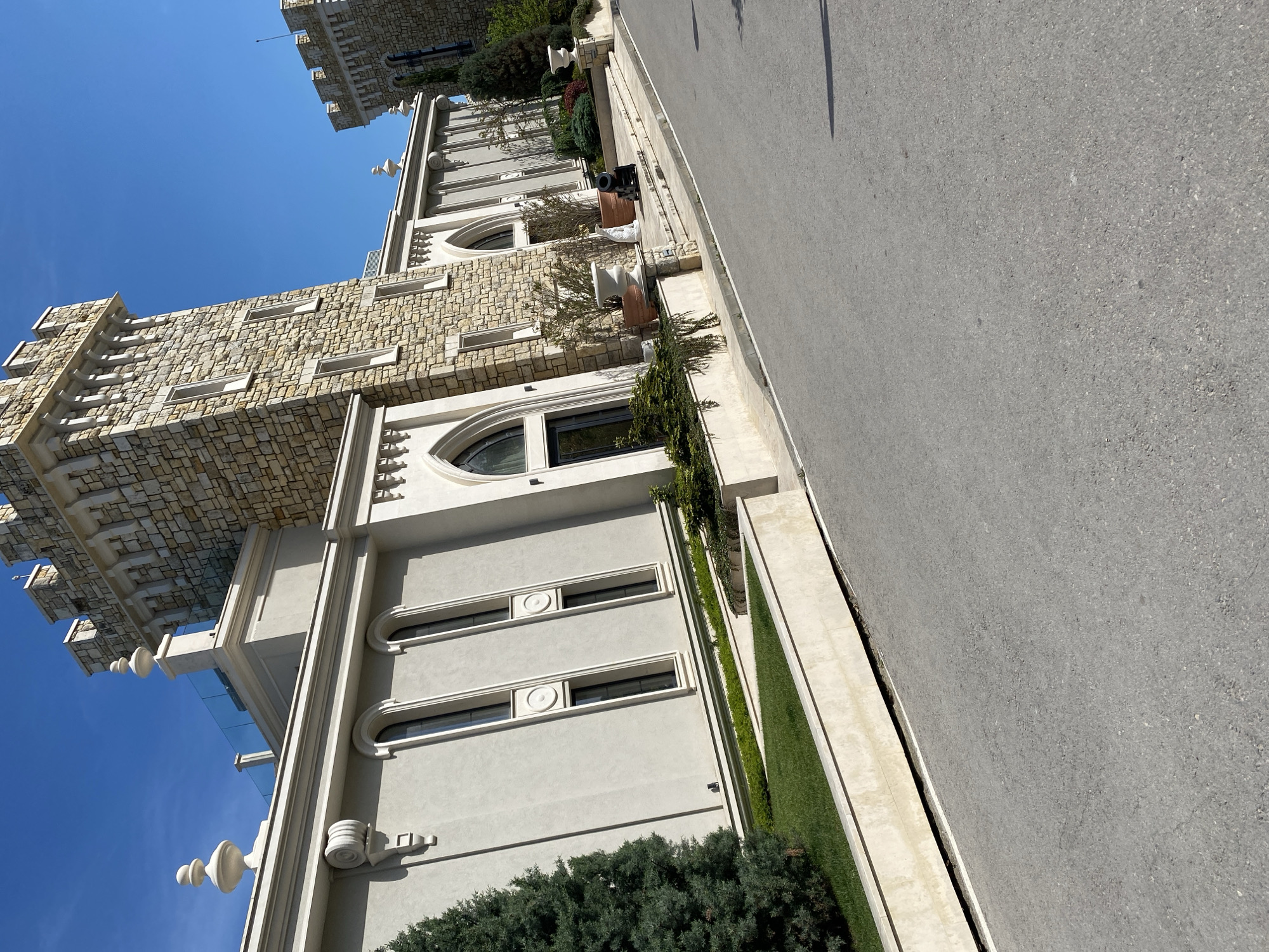 The image shows the exterior of a building in the city of Unknown, Unknown. The building features a combination of light-colored stone and stucco materials. It has architectural details such as arched windows and decorative trim. The building is adjacent to a paved road or sidewalk. There are green plants, including shrubs and grass, along the base of the building. The sky is clear and blue. A portion of another building can be seen in the distance.