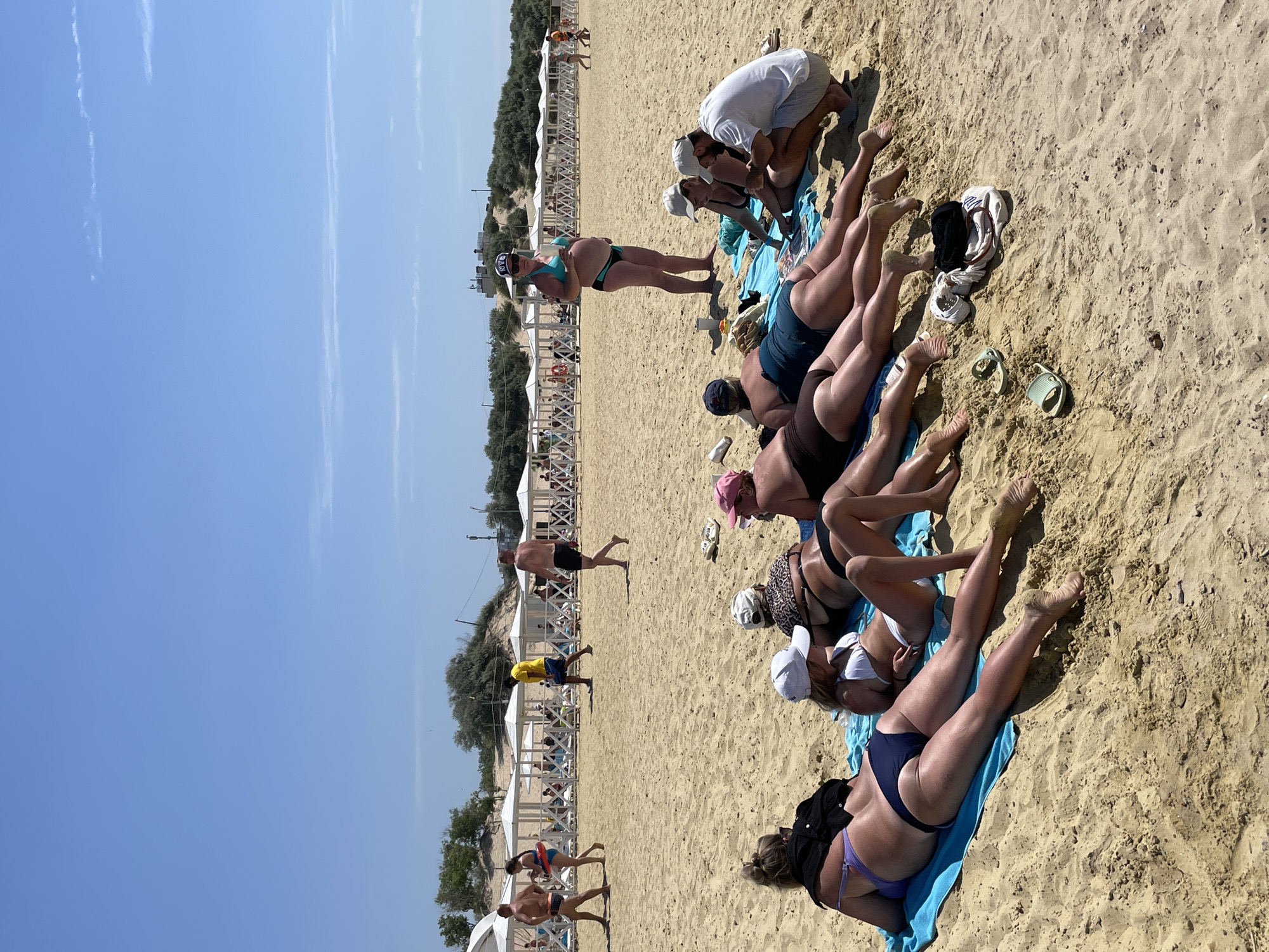 The image depicts a sunny day at a beach, likely in Anapa, Russia, given the presence of beach umbrellas and a sandy coastline. In the foreground, several people are lying on towels on the sand, some in bikinis and swim trunks, soaking up the sun. Their bodies are partially visible as they recline. In the middle ground, more people are scattered across the beach, with some walking and others sitting under white beach umbrellas. A person in a yellow t-shirt walks in the distance, while a pregnant woman in a turquoise bikini stands near the umbrellas. The background reveals a line of beach tents and lush green vegetation beyond the sand. The overall scene is one of a typical summer day at a crowded beach.