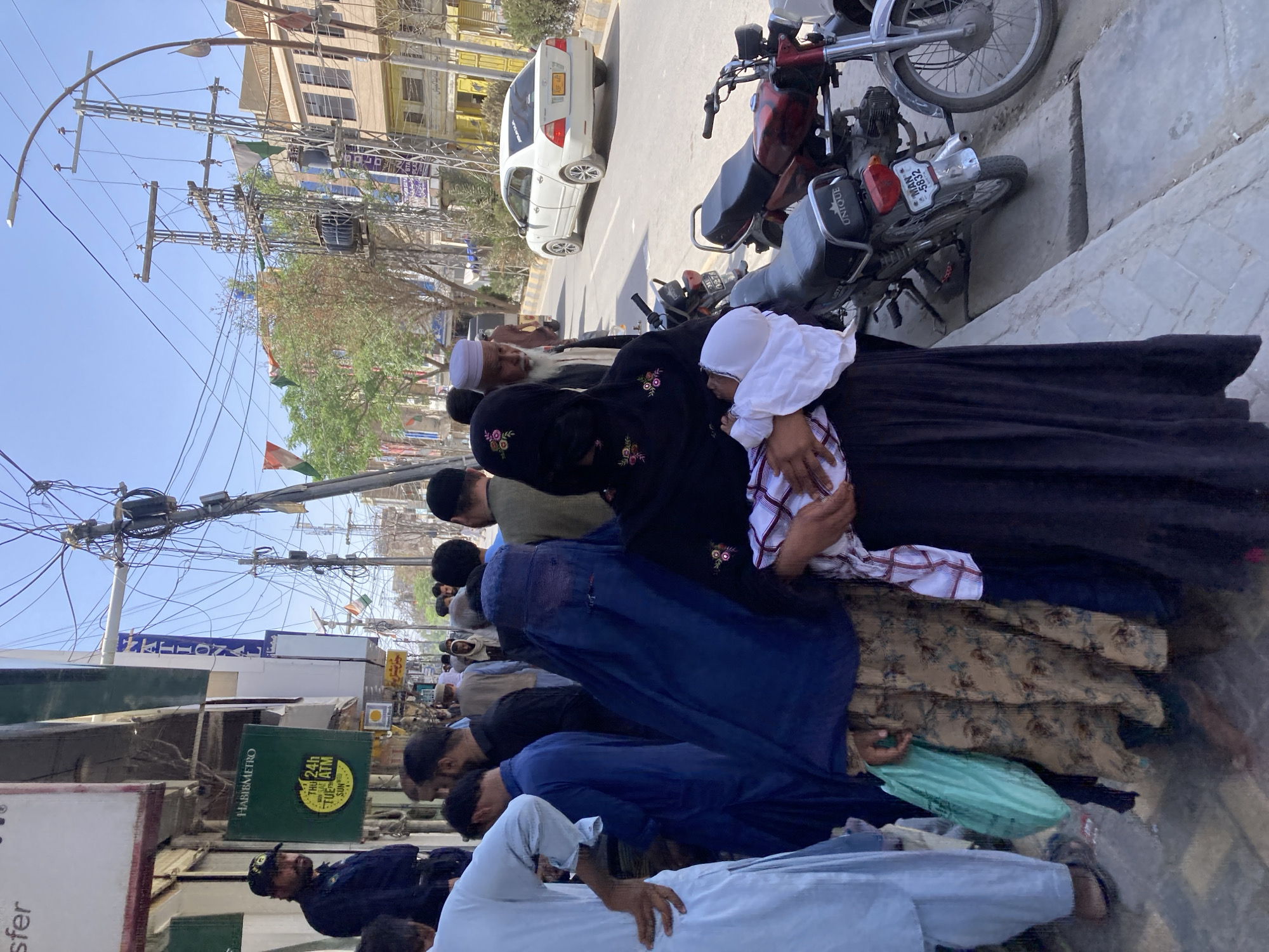 In Quetta City Tehsil, Pakistan, a group of people are prostrating on a city sidewalk. Several are men in traditional Muslim clothing. Several women wearing black abayas are present, one of whom is holding a child in white. The group is praying in a line extending along the sidewalk. Motorcycles are parked beside the praying group, and a white car is visible in the street. Power lines and buildings can be seen overhead and in the background. A sign for Habib Metro bank is visible. Flags of Pakistan are hanging above.