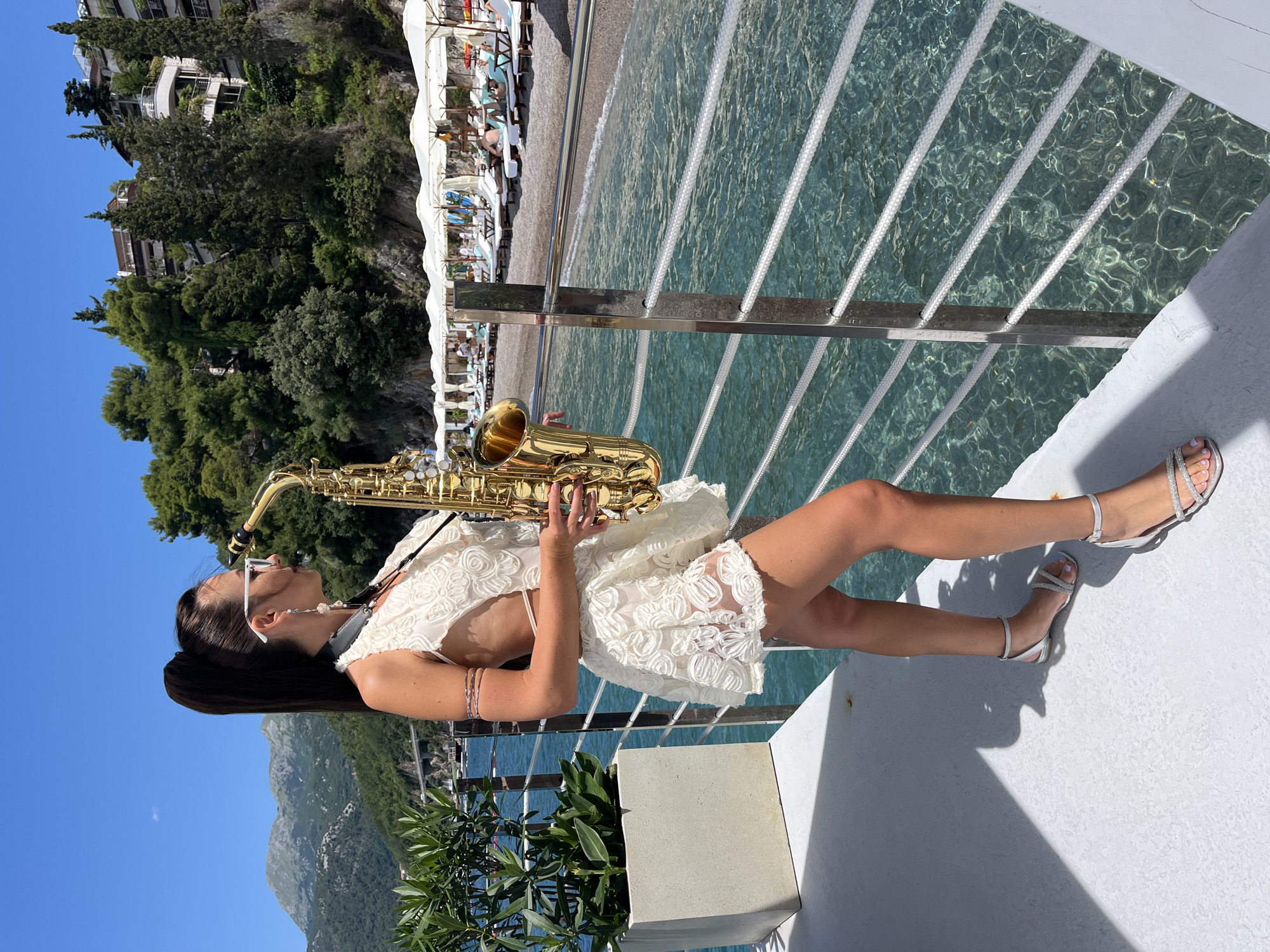 A woman stands on a white deck by a railing, holding a saxophone. She wears a white, lace-trimmed dress and silver sandals. The background features a clear blue sea, a pebble beach with rows of lounge chairs and umbrellas, lush green trees, and buildings nestled into a hillside. Mountains are visible in the distance under a clear blue sky. The scene is set in Budva, Montenegro.