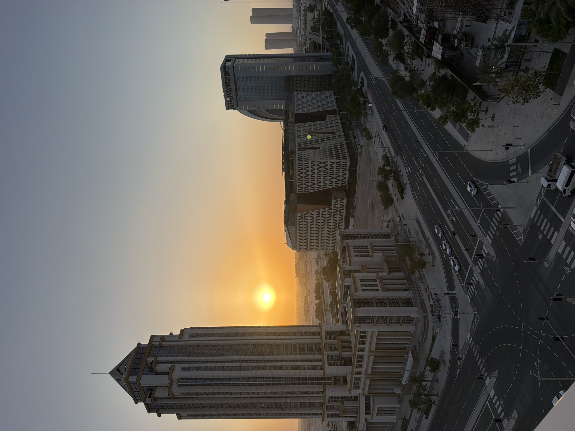 A high-angle, medium shot captures an urban landscape at sunset in an unknown city. The sun, a bright orb of light, is positioned to the left of the frame, casting a warm, orange glow across the sky and illuminating the scene. In the foreground, a prominent skyscraper with a tiered design and a pointed roof dominates the left side of the image. Its facade is a mix of light-colored stone and glass windows. Adjacent to it is a lower, rectangular building with a patterned facade. Further into the midground, a cluster of modern buildings with distinctive architectural features stand. One building has a curved facade, while another is characterized by a geometric pattern. In the distance, several taller, slender buildings are visible, suggesting a developed urban area. A multi-lane road with traffic markings runs through the scene, with a few cars visible. Sidewalks, trees, and streetlights line the road. The overall environment appears to be a modern district with ongoing construction visible on the right side of the frame, indicated by scaffolding and machinery. The sky is clear, transitioning from a pale blue at the top to a warm orange near the horizon.