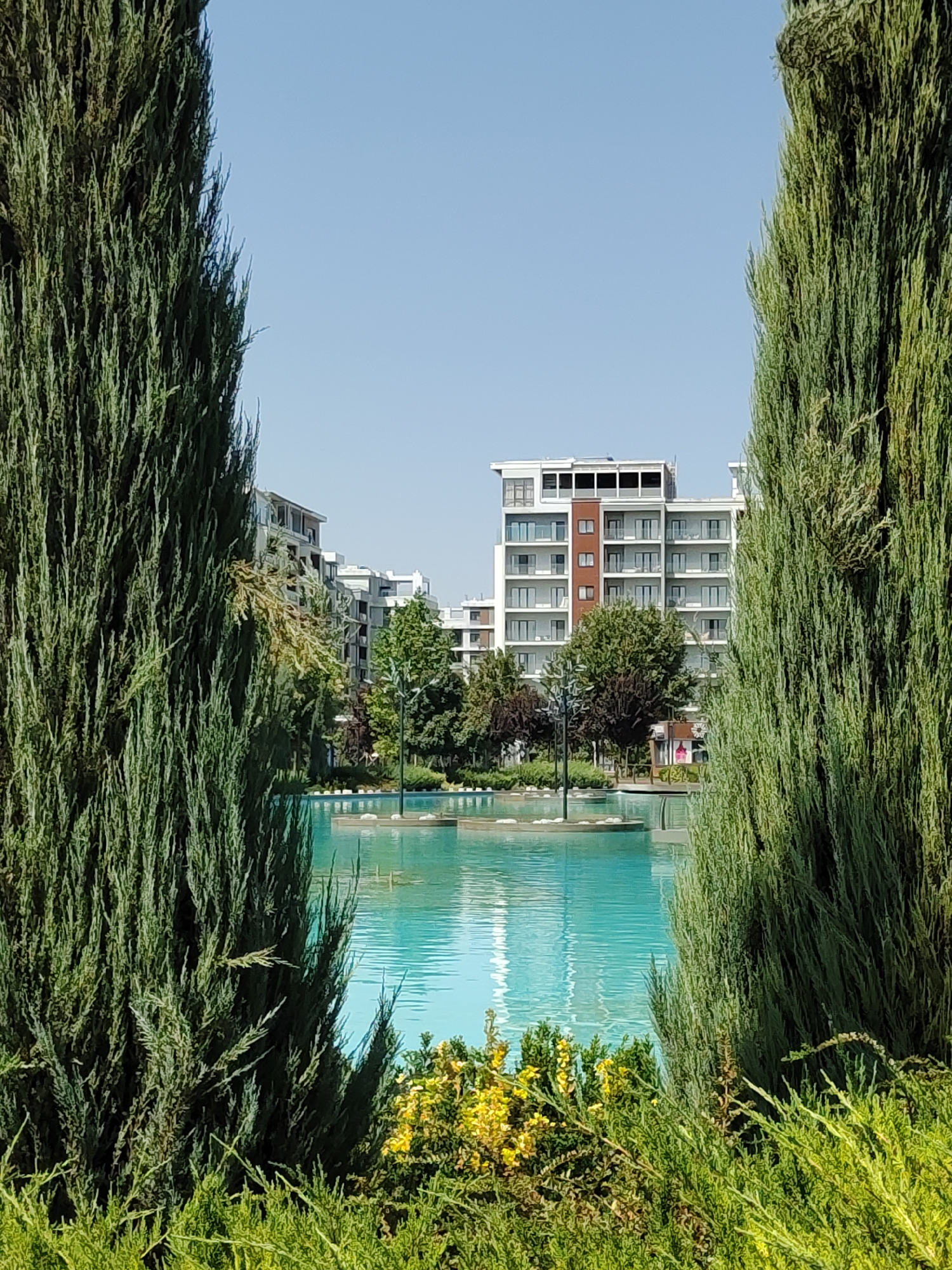 The image depicts a modern residential complex situated in the city of Tashkent, Uzbekistan. In the foreground, two tall, mature cypress trees frame a view of a large, turquoise swimming pool or decorative pond. The water is calm with gentle ripples and reflects the bright, clear blue sky above. Several small, circular islands are visible within the pool, some featuring decorative elements and a lamppost.
Beyond the pool, a multi-story building with a contemporary architectural design stands prominently. It features a light-colored facade with numerous balconies and large windows. The building's design suggests a modern apartment complex. Surrounding the building are various types of trees and greenery, indicating a well-maintained landscape. A lamppost is also visible in the midground. The overall scene conveys a sense of urban tranquility and modern living within a green environment. There are no people visible in the image.