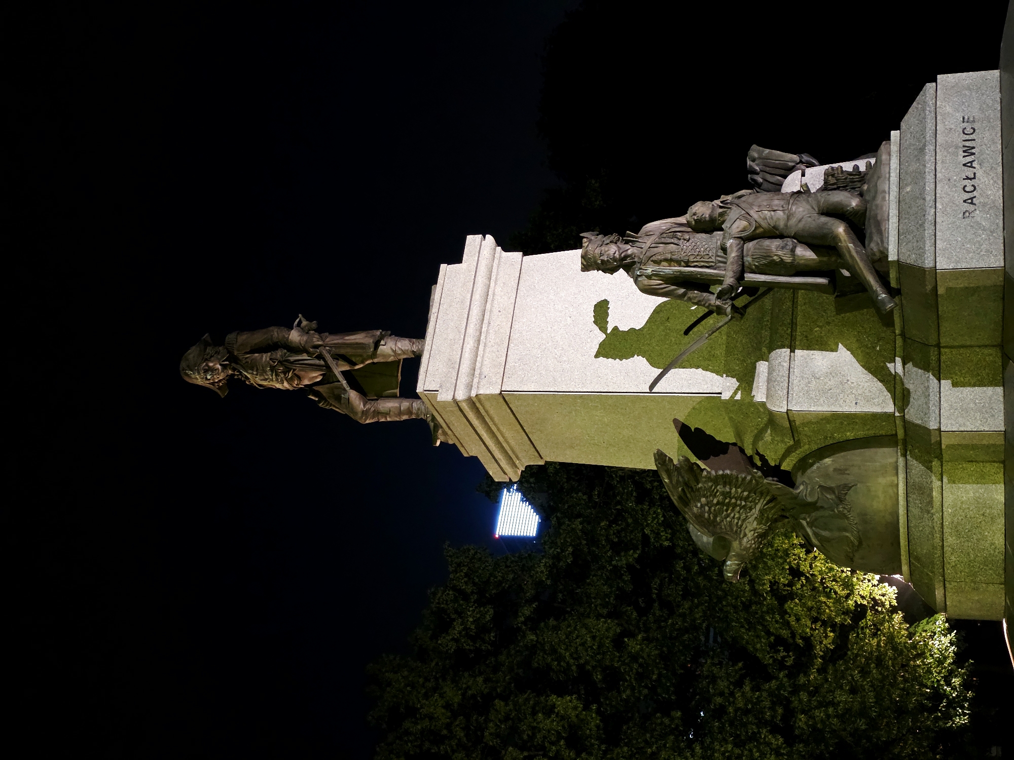 The image shows a nighttime view of the Monument to the Heroes of Racławice in Warsaw, Poland. The monument features a central pedestal topped by a statue of a standing man in military attire, holding a sword. At the base of the pedestal, there are two additional bronze figures: one standing man with a sword and a headdress, and a kneeling man. To the left of the pedestal, there is a bronze sculpture of an eagle. The monument is illuminated, casting shadows on its surfaces. Behind the monument, a dark tree with some illuminated foliage is visible to the left, and a distant building with a glowing facade is visible in the upper center. The word "RACŁAWICE" is inscribed on the base of the monument.