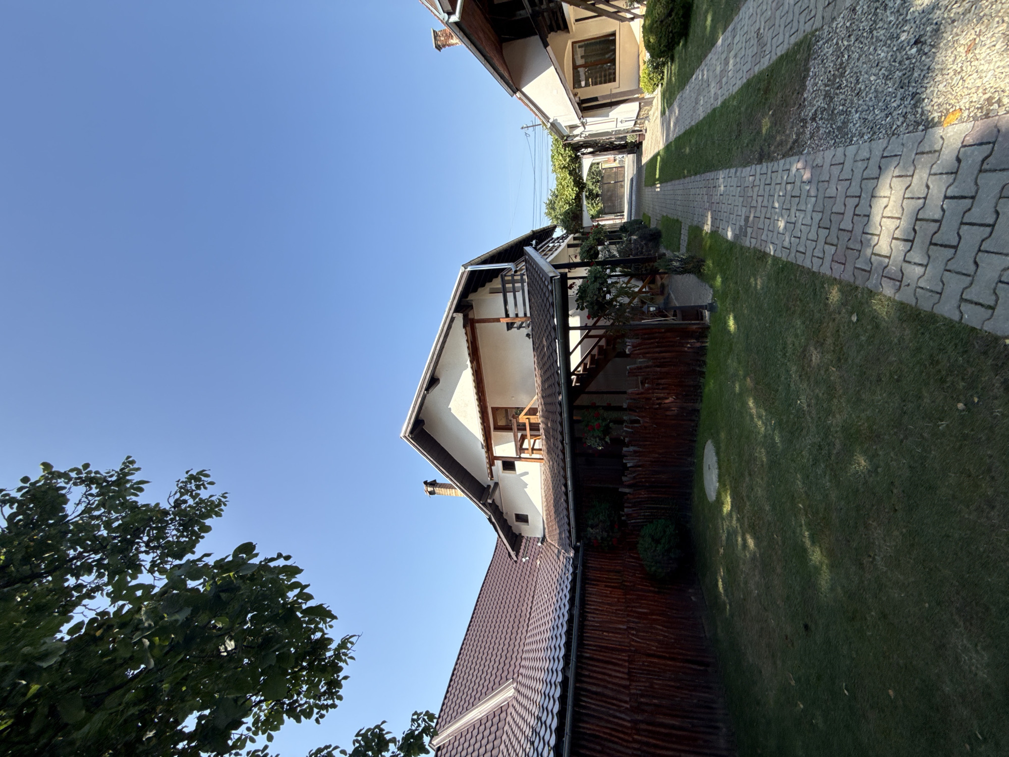 A photograph taken on a sunny day in Cârțișoara, Romania, shows a residential area with two houses and a paved pathway. The house on the left has a brown, tiled roof and a rustic wooden fence. The house on the right is lighter in color and also has a tiled roof. Both houses appear to be two-story structures with balconies and outdoor stairs. A cobblestone pathway runs between the houses, leading towards a large archway in the distance. The pathway is bordered by green grass and small bushes. Trees with green leaves are visible, casting shadows on the ground. The sky is clear and blue, indicating fair weather. There are no people or animals visible in the image. The scene depicts a peaceful, well-maintained residential property.