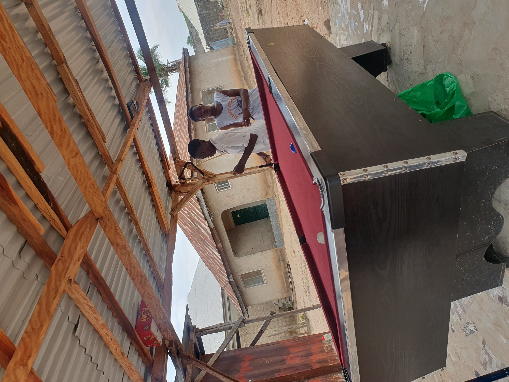 In the city of Lokoja, Nigeria, two young males are standing by a dark brown pool table with a red felt surface. The pool table is positioned outdoors under a rustic wooden structure with a corrugated metal roof, suggesting an open-air establishment or recreational area. One individual, wearing a white shirt, holds a pool cue. The other, in a sleeveless white t-shirt, stands with his hands clasped. A blue pool ball is visible on the felt. Behind them, a light-colored building with a green door and rusted corrugated iron roofing is visible, typical of local architecture. The ground is sandy and uneven. A green plastic bag is situated under the pool table. The overall environment appears to be a casual, possibly informal, outdoor setting for games.