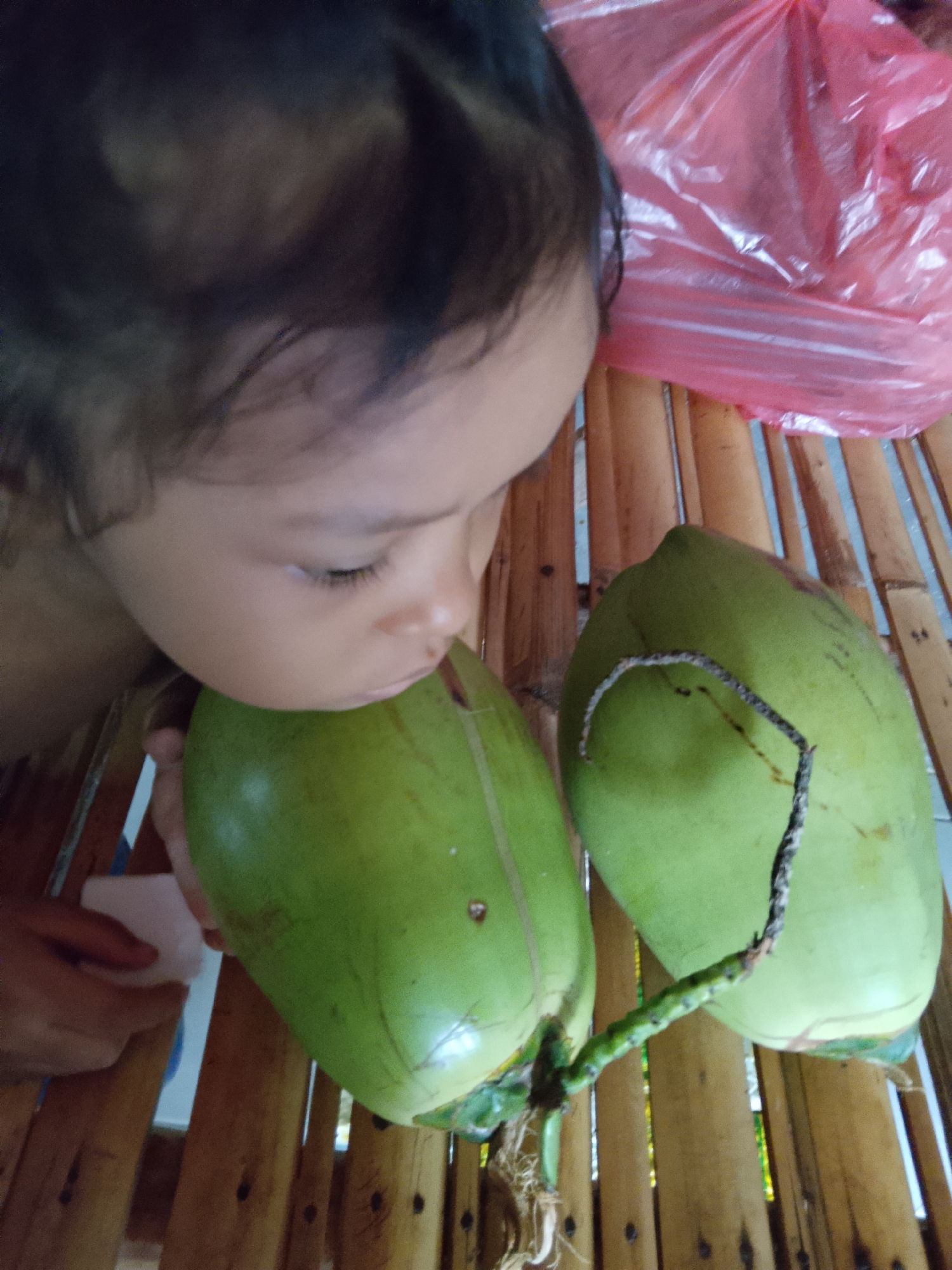A young child with dark hair is leaning towards two green coconuts. The child's head is angled down, with their eyes closed or looking down at the coconuts. One coconut is in the foreground, with the child's face close to it. The second coconut is to the right and slightly behind the first. Both coconuts have green stems attached. The background consists of a slatted surface, likely made of bamboo, and a crumpled pink plastic bag. The image appears to be taken indoors, with the lighting suggesting it is daytime. The location is identified as Merbopuntong, Indonesia.