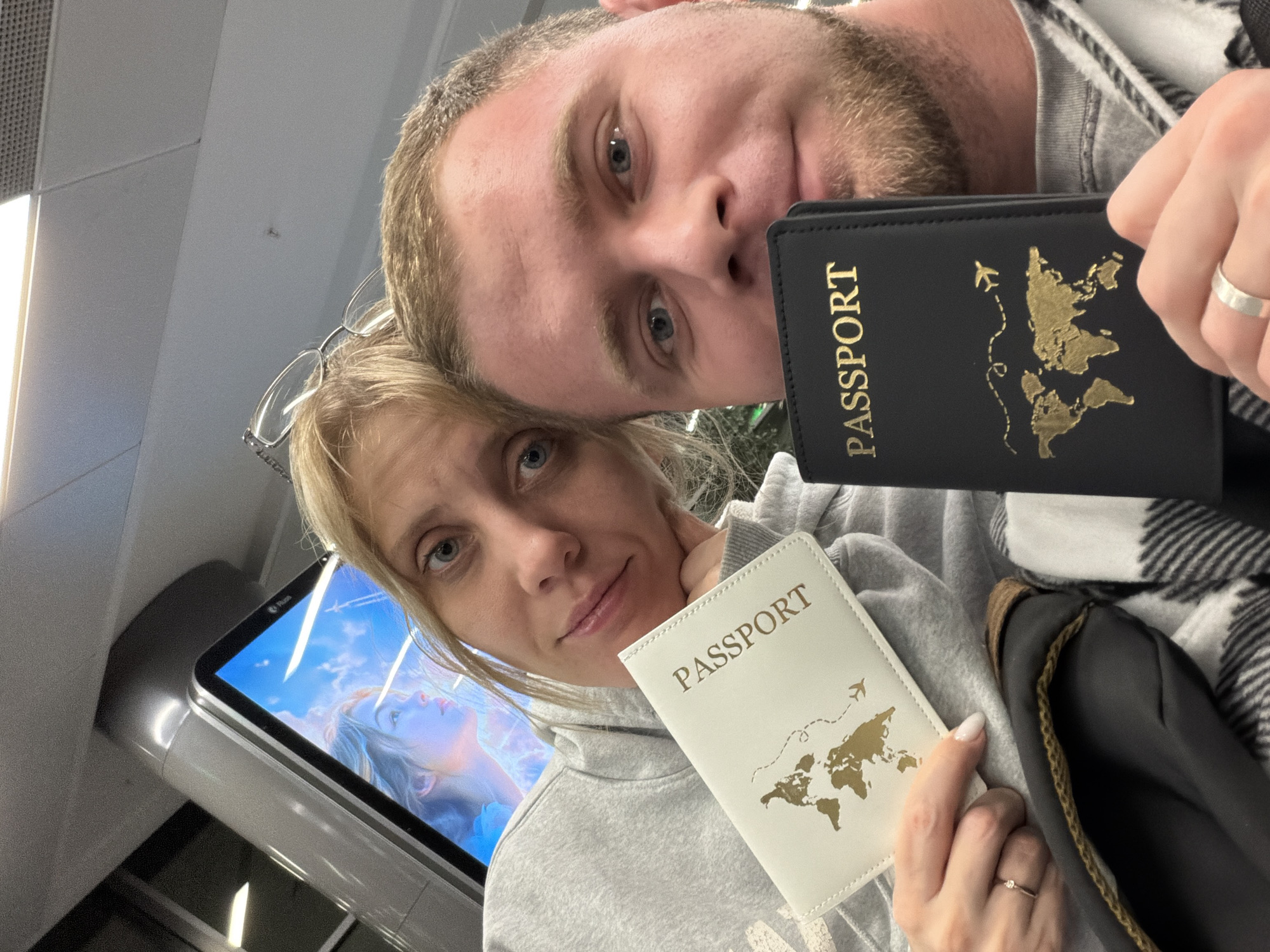 The scene depicts two individuals, one female and one male, captured in an indoor selfie-style photograph. The scene is an indoor setting in the city of Boldino, Russia.
The female, positioned on the left, has blonde hair, blue eyes, and wears glasses pushed onto the top of her head. She is wearing a grey hooded sweatshirt and holds a white passport cover in her left hand, featuring the word "PASSPORT" and a gold world map design. A thin silver ring with a small stone is visible on her left ring finger. Her chin rests on her right hand.
The male, positioned on the right and partially obscured by the female, has light brown hair, blue eyes, and light brown facial stubble. He wears a grey garment and a black and white plaid pattern, possibly a blanket or scarf, is draped over him. He holds a black passport cover in his right hand, which also displays "PASSPORT" and a gold world map design, complete with an airplane icon and a dashed flight path. A plain silver band is visible on his left ring finger.
In the background, a digital display screen shows a partial image of a person against a blue, cloudy sky. Overhead, a standard drop ceiling with recessed lighting fixtures is visible, along with a grey structural support column. A dark bag or object is partially visible near the female's left arm.