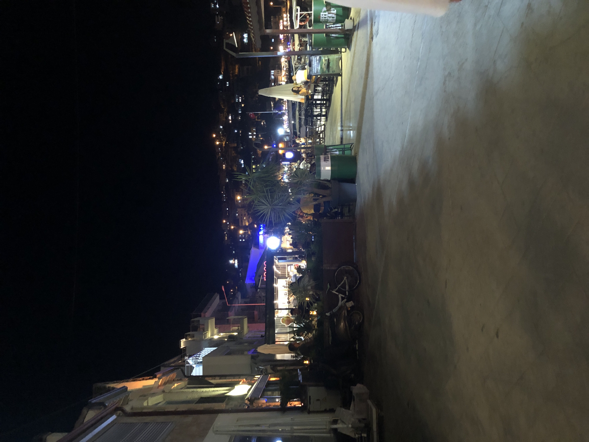 The image captures a lively night scene in Karaburun, Turkey, likely a coastal town. The foreground is dominated by a paved walkway, reflecting the ambient light from the establishments lining the street. To the left, a row of buildings with illuminated windows and balconies suggest commercial and residential spaces, with a warm orange glow emanating from some of them. Further back, a cluster of outdoor seating areas for restaurants and cafes is visible. People are seated at tables, some engaged in conversation, while others appear to be dining or enjoying drinks. Several large potted plants, including what appears to be a yucca, add greenery to the scene, interspersed with decorative elements like candles on tables. A scooter and a bicycle are parked near the seating area, suggesting modes of local transportation.
Beyond the immediate seating areas, the scene opens up to a harbor or waterfront. Boats are docked, their lights twinkling on the water. The background is a hillside densely populated with houses, their lights forming a sparkling tapestry against the dark night sky, characteristic of a Mediterranean coastal town. Streetlights and building lights illuminate the area, creating a festive atmosphere. The overall impression is one of a popular evening destination, with a vibrant social scene set against a beautiful nighttime landscape.