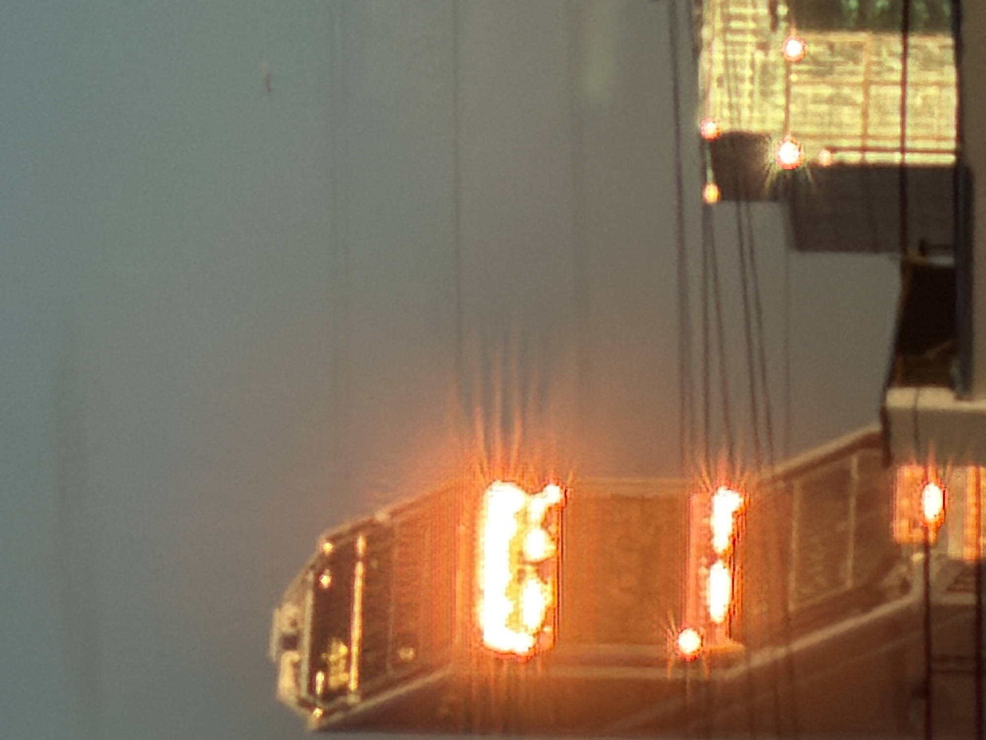 The image shows a view of the Khimki, Russia skyline, likely at dusk or dawn, with buildings and a structure resembling a communication tower. The tower, prominently featured on the left, is illuminated with bright orange and white lights, suggesting it is actively in use or decorative. The lights create a halo effect due to the image's resolution. Behind the tower, a portion of another building, a tall rectangular structure with horizontal and vertical lines of light, is visible on the right side of the frame. The sky is a muted, dusky blue-gray, with thin, horizontal lines, possibly power lines, crossing the scene. The overall impression is one of an urban landscape at low light, with man-made structures illuminated against the fading daylight.