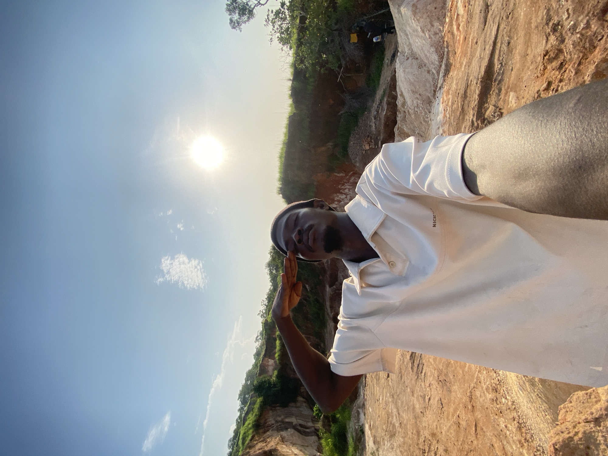 A young Black man, wearing a light-colored collared shirt and a dark beanie, salutes while standing in a dry, rocky, and dusty outdoor environment. Behind him, a natural landscape features hills with green vegetation and exposed reddish-brown earth. The sun is visible in the bright blue sky, casting light and shadows on the scene. The image appears to have been taken in a quarry or a similar geological site. The location is identified as Ufu Esioni, Nigeria.