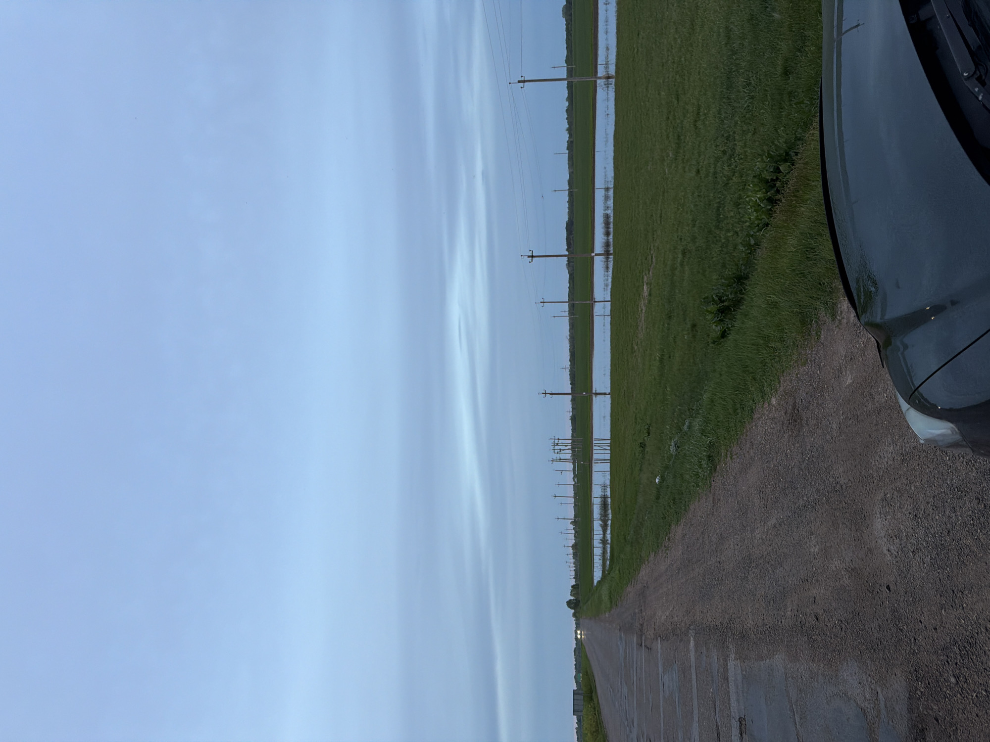 The image shows a straight, narrow dirt road in Azyakul', Russia. Power lines run along the roadside through a flooded, grassy field. A portion of a dark-colored car is visible in the right foreground. The sky is overcast with light blue tones. The landscape appears rural.