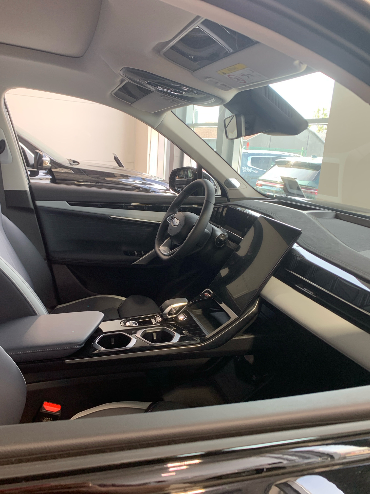 An interior view of a black Geely electric vehicle showcases the driver's side and dashboard. The seats are upholstered in a two-tone grey and black material, with visible stitching. The dashboard features a large, modern touchscreen display angled towards the driver, a sleek steering wheel with integrated controls, and prominent air vents. The center console includes a gear selector, cup holders, and various buttons and switches. The overall impression is one of a contemporary and high-tech vehicle interior. The image appears to have been taken at a car dealership or showroom. The presence of other vehicles in the background, visible through the windows, supports this interpretation. The ambient lighting suggests it is daytime. The cropped images provide closer views of the ceiling console, rearview mirror assembly, door panel, and the prominent touchscreen.
