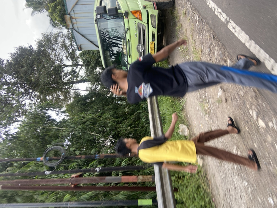 Two young men are walking along a paved road in Cipatat, Indonesia. The men are walking in the same direction, with the man in the yellow shirt walking slightly ahead of the man in the black shirt. The man in the yellow shirt is wearing brown pants and sandals. The man in the black shirt is wearing grey pants and sandals. Both men are walking casually. There is a large green truck parked on the side of the road to the right of the men. There are numerous poles, possibly for power lines, on the left side of the road. The road is bordered by a guard rail and a dense patch of green trees and bushes.
