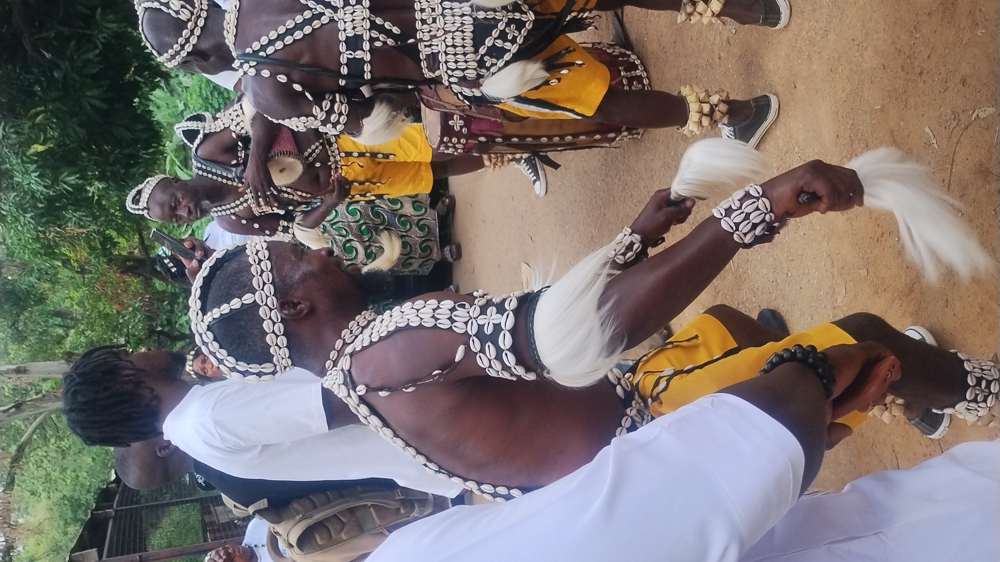 The image shows a group of men in traditional attire, seemingly participating in a cultural ceremony. They are adorned with necklaces and headdresses made of cowrie shells. Some are wearing yellow shorts and elaborate beaded accessories. One man in the foreground is holding a white feathered object and is adorned with cowrie shells on his arms and legs. Another man in the background is holding a small drum. The setting appears to be outdoors, with a sandy ground and vegetation visible in the background, suggesting a natural or semi-urban environment. The attire and context suggest a cultural event possibly taking place in Jos, Nigeria, known for its rich cultural heritage and diverse ethnic groups.