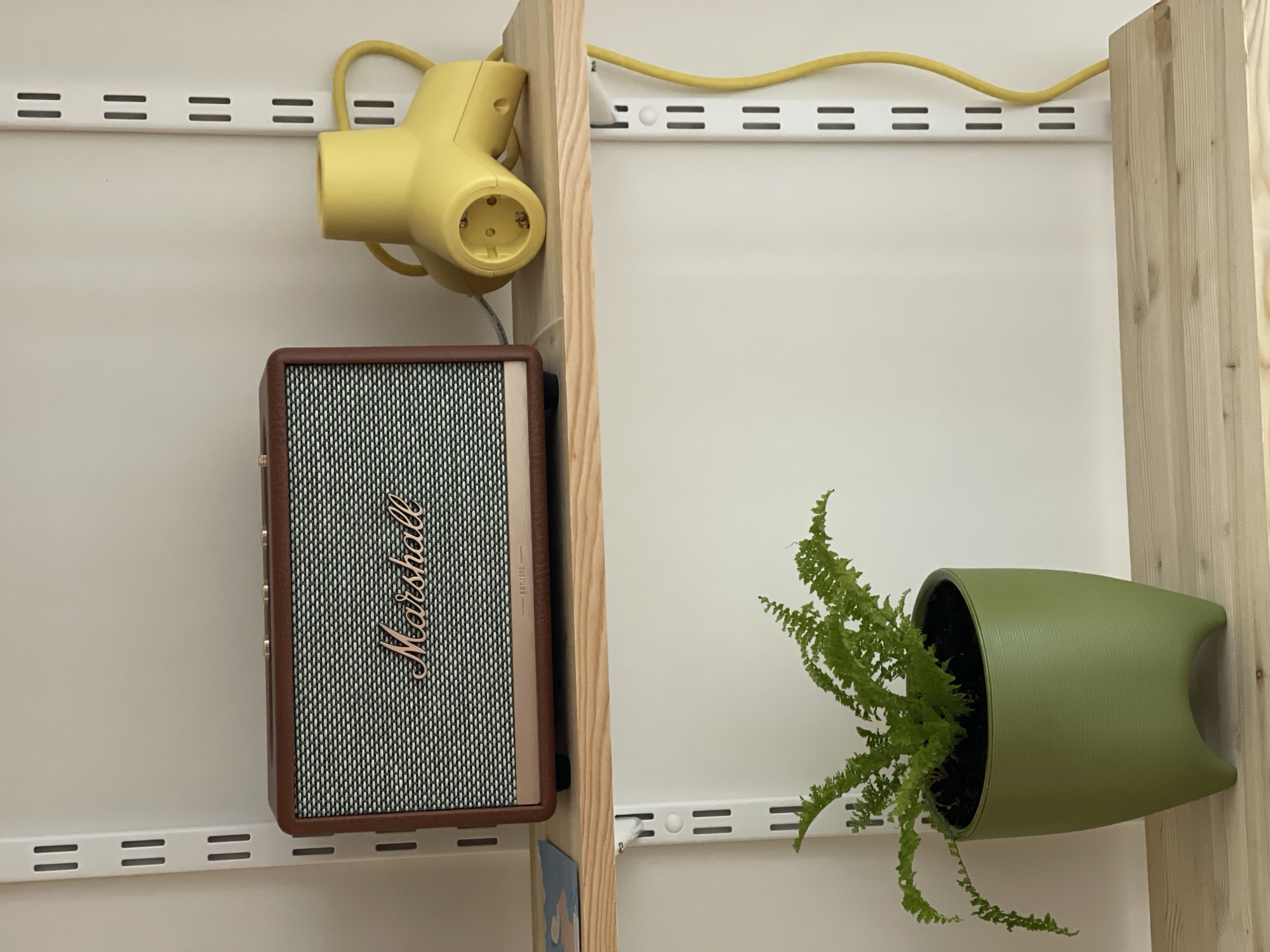 The image shows a section of a white wall with a shelving system. Attached to the shelving are a yellow power strip, a Marshall speaker, and a green planter with a fern. The shelving system has horizontal metal tracks with evenly spaced slots. A wooden beam runs vertically alongside the shelving on either side of the image. The objects are attached to the shelving system using brackets or hooks. The power strip has a yellow cord that extends across the top of the image. The Marshall speaker is brown and black. The planter is a solid green color and is oriented horizontally. The fern is lush and green. There is no indication that the image was taken in Izhevsk, Russia.