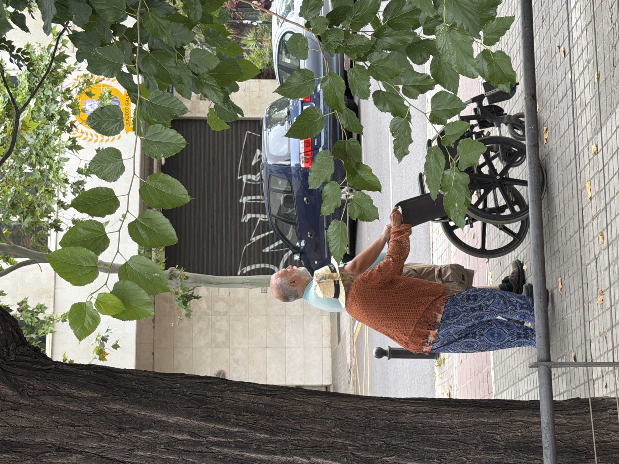 An elderly couple is walking down a sidewalk in an urban environment. The man, wearing a light blue polo shirt and tan shorts, is standing behind the woman, who is wearing a patterned orange knitted sweater and blue patterned pants, and pushing a wheelchair. The wheelchair is black with large spoked wheels. The sidewalk is paved with square tiles and bordered by a metal railing. Behind them, a dark blue minivan and a silver car are parked on the street, which has yellow lane markings. A large tree trunk is visible on the left side of the frame, and green leaves from a tree canopy hang over the scene. A building with a beige tiled facade and a closed garage door is in the background, along with a circular logo featuring a profile of a person.