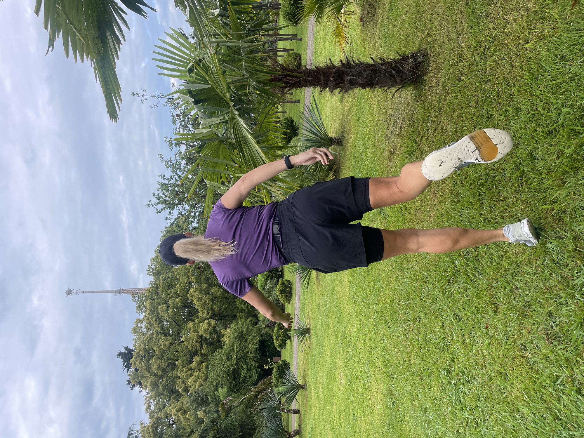 A woman stands on a green lawn, her back to the viewer. She wears a purple shirt, black shorts, and athletic shoes, and a black hat. Her blonde hair is long. She extends one leg behind her and her arms are at her sides. The scene includes palm trees and other greenery, with a tower visible in the distance. The location is the Arboretum in Sochi, Russia.