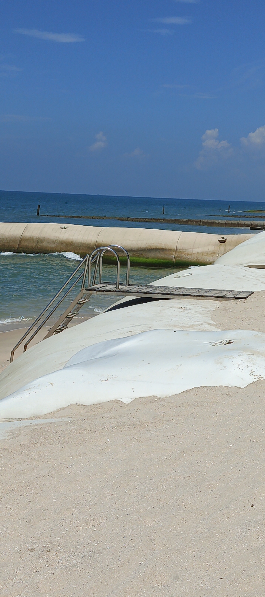 The image depicts a section of a beach in Phu Thuy Ward, Vietnam, characterized by a calm sea under a bright blue sky with scattered white clouds. In the foreground, a sandy beach leads to a large, white, smooth mound, likely a sandbag or geotextile structure used for coastal defense. To the left, a metal ladder with curved handrails ascends from the water onto a wooden platform, suggesting a diving board or access point to a submerged structure. Beyond the immediate shoreline, a long, cylindrical object, possibly another large sandbag or barrier, extends horizontally across the water, partially submerged and covered in green algae. Further out at sea, a series of vertical poles are visible, some of which appear to be part of another long, low barrier structure. The water is a clear, light blue-green, with gentle waves lapping at the shore. The scene is devoid of any people.