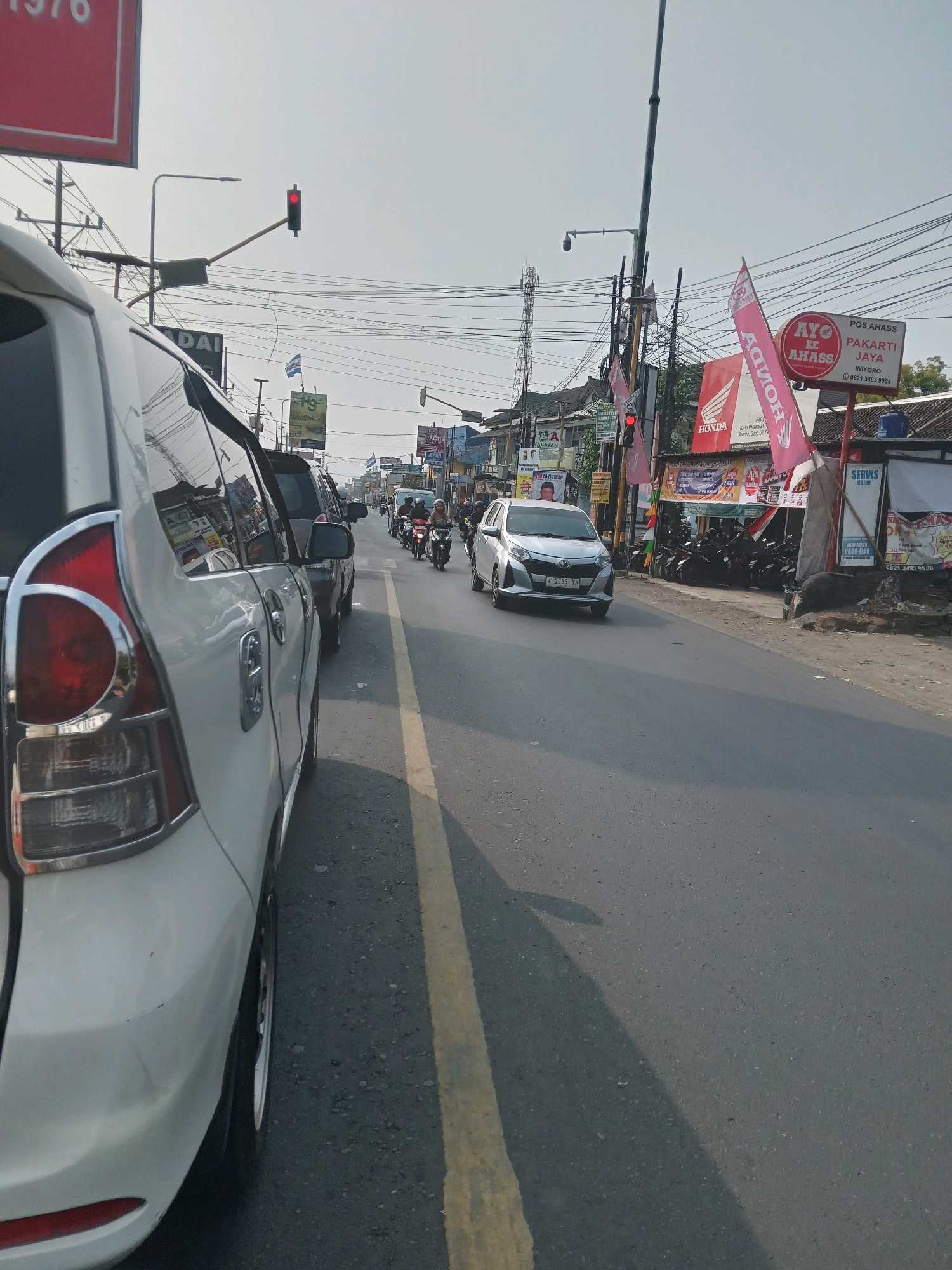 The image depicts a street scene in Banguntapan, Indonesia, on a clear day. The foreground features the rear of a white car, showing its taillight and wheel. The street is paved with asphalt and marked with a yellow lane divider. Several other vehicles, including cars and motorcycles, are visible on the road, suggesting moderate traffic.
Along the right side of the street, there are various shops and businesses. Prominent signage includes "AYO KE AHASS," indicating a Honda service center, with flags bearing the Honda logo. Other businesses display advertising for services and products. Power lines crisscross overhead, supported by poles. A traffic light is visible in the distance, showing a red signal. The overall environment suggests a commercial area within the town. People are seen riding motorcycles, and some individuals are present near the shops. The sky is clear and bright.